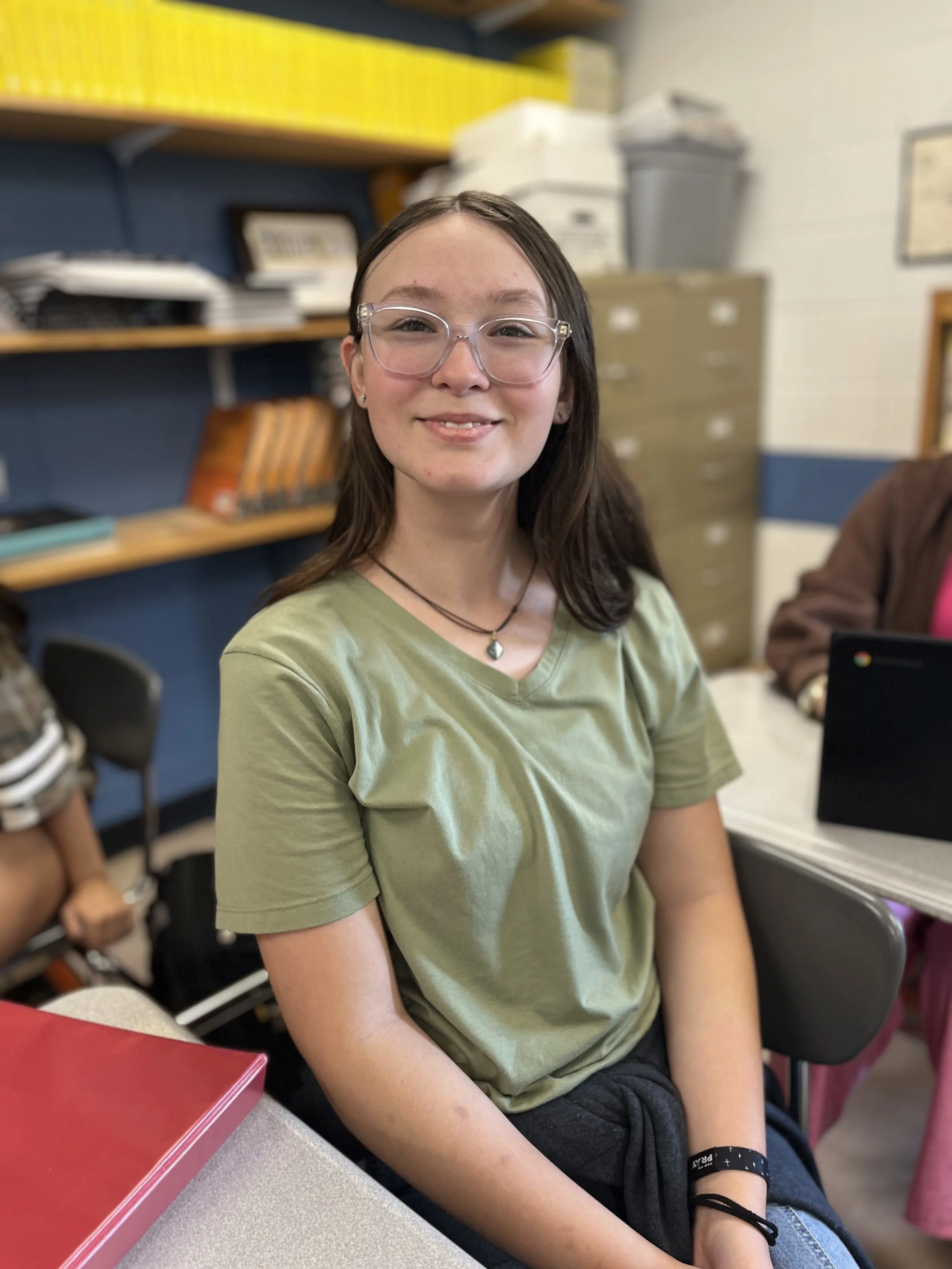 A young woman smiling, sitting in a classroom, with two other students blurred in the background.