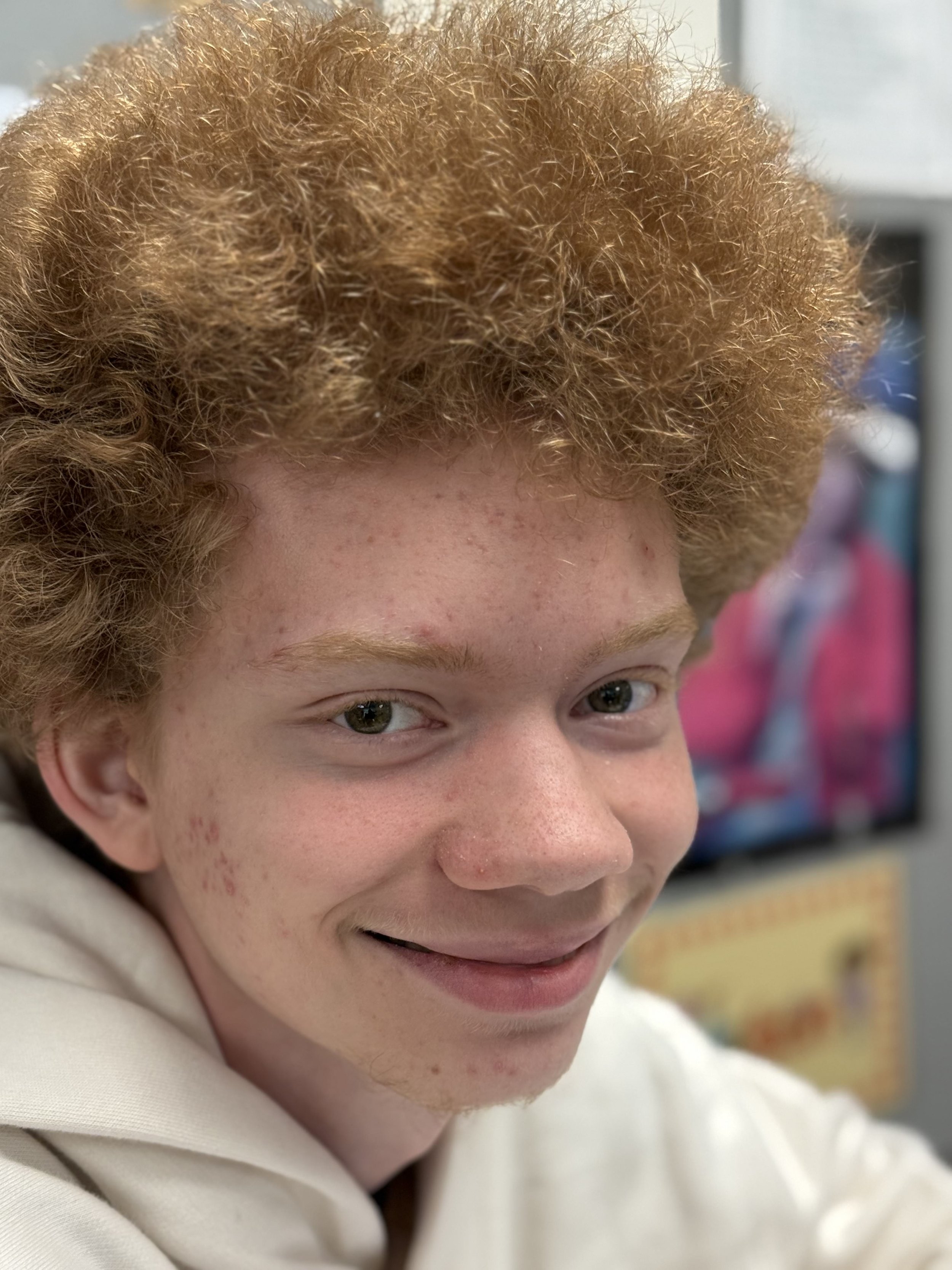A young person with curly red hair and light skin, smiling in a classroom with a colorful poster or book in the background.