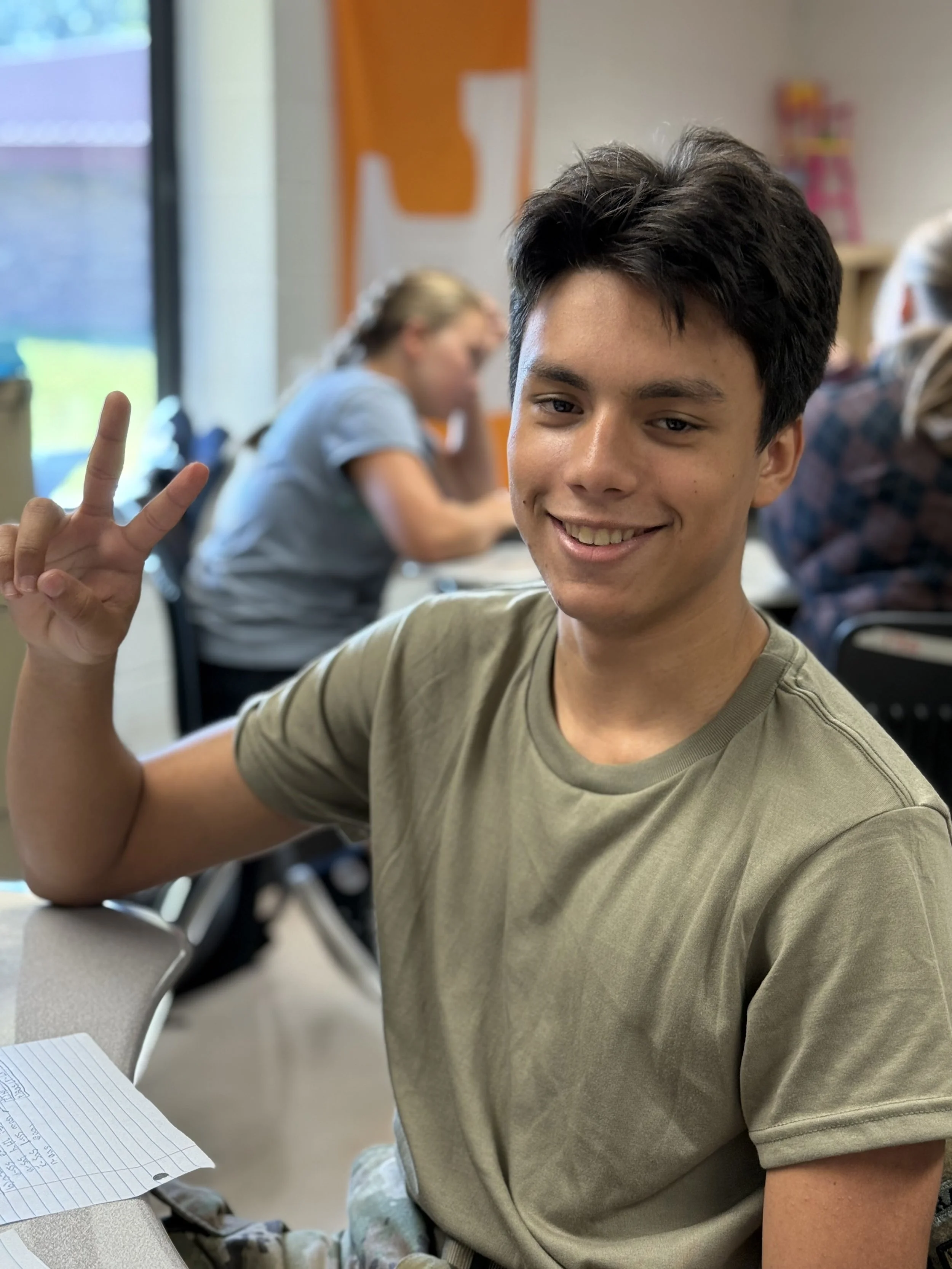 A young man smiling and making a peace sign with his hand in a classroom setting, with other students in the background.