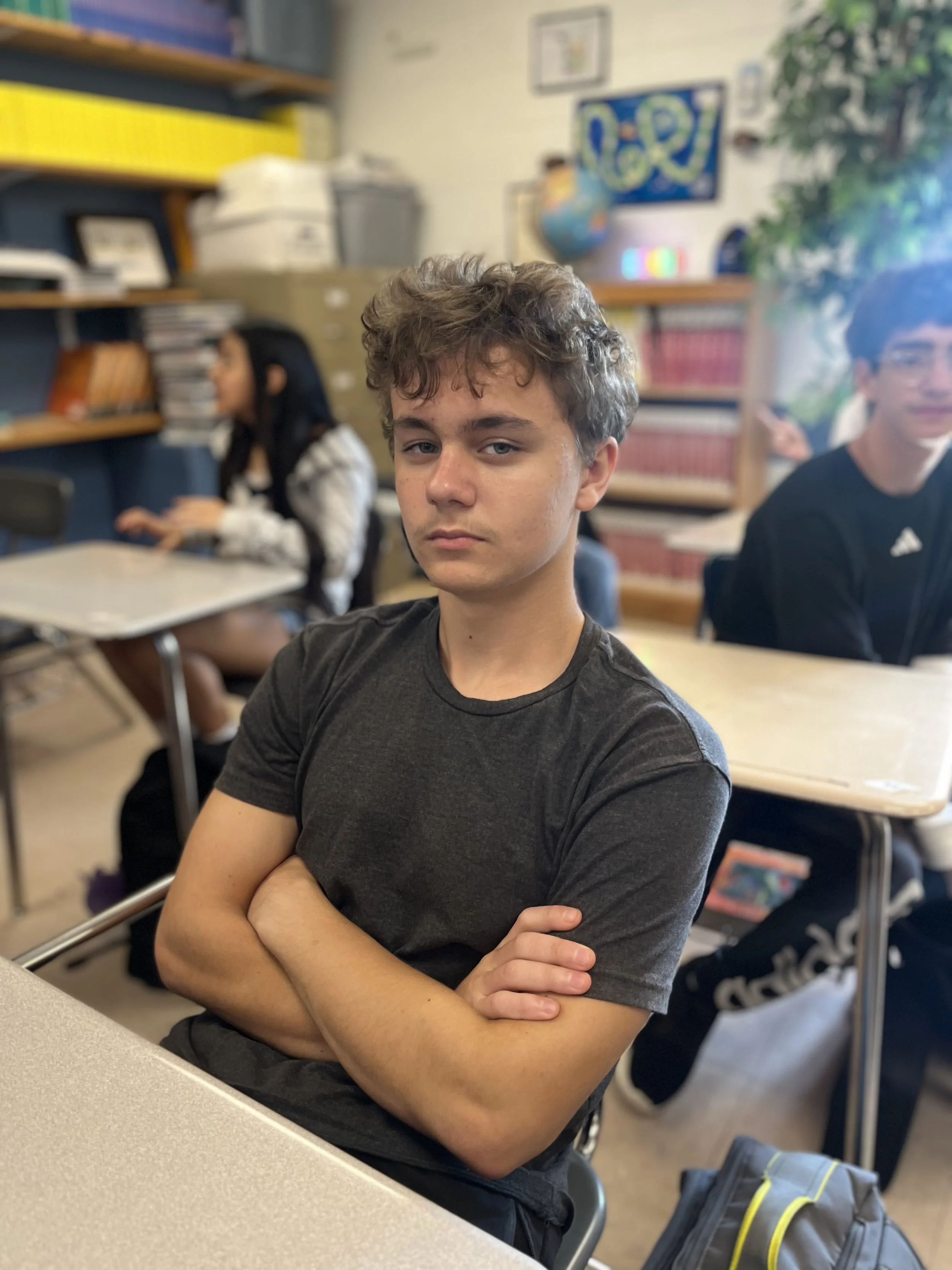 A teenage boy with light skin, brown hair, and a slight smile, wearing a blue T-shirt with white text and graphics, standing indoors.