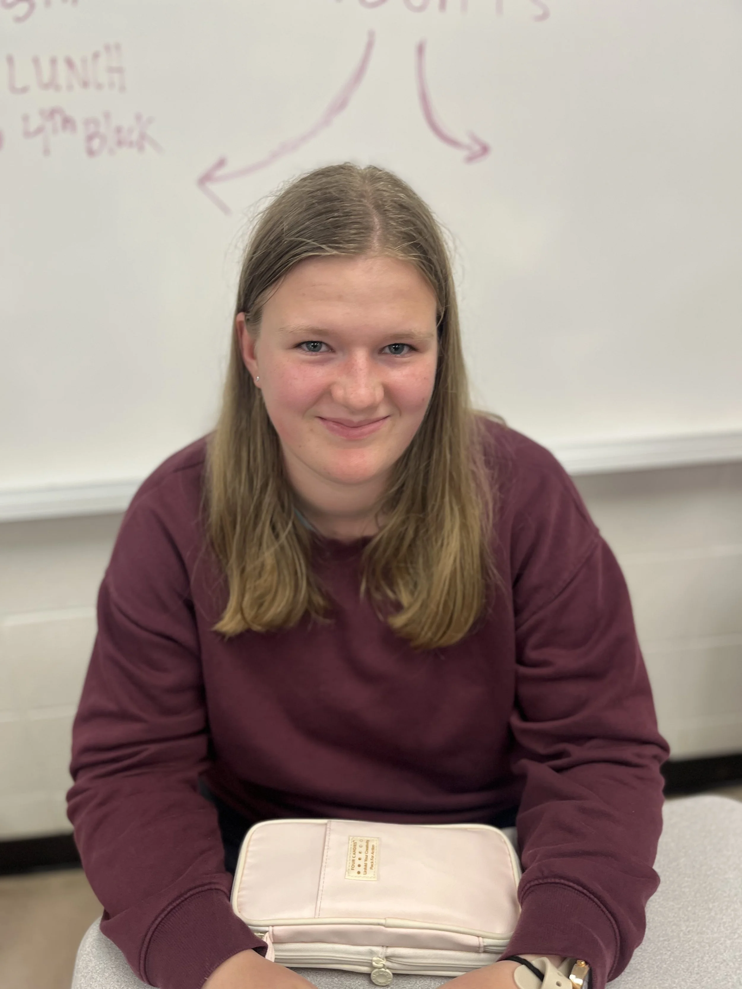 A smiling young boy with short light brown hair, wearing a dark gray T-shirt, sitting in a classroom or office environment with shelves, books, and a desk in the background.