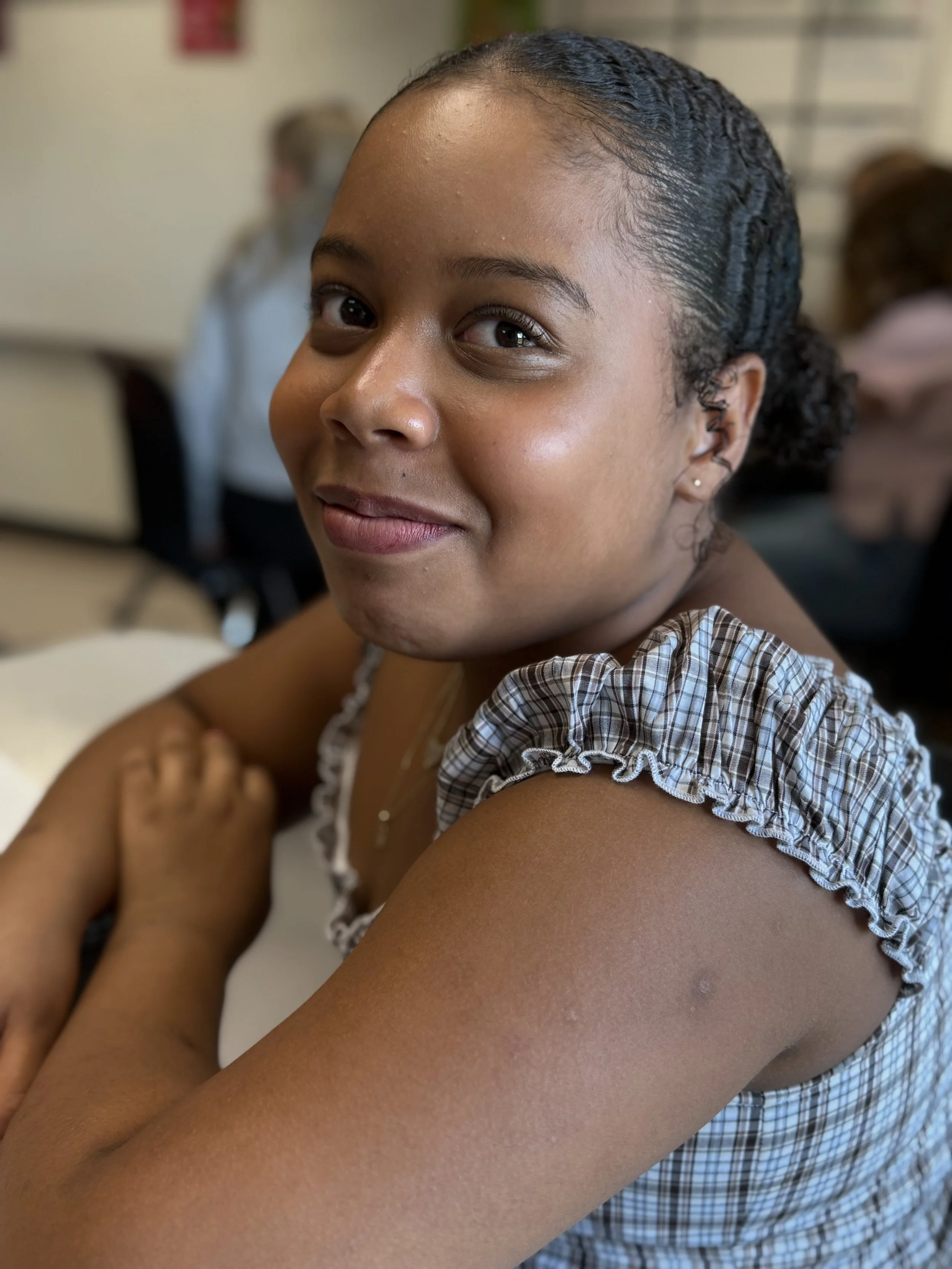 A young girl with a warm smile, sitting indoors, with her arms crossed on a table. She has dark braided hair and is wearing a plaid dress with ruffled sleeves, with a background that appears to be a classroom or similar setting.