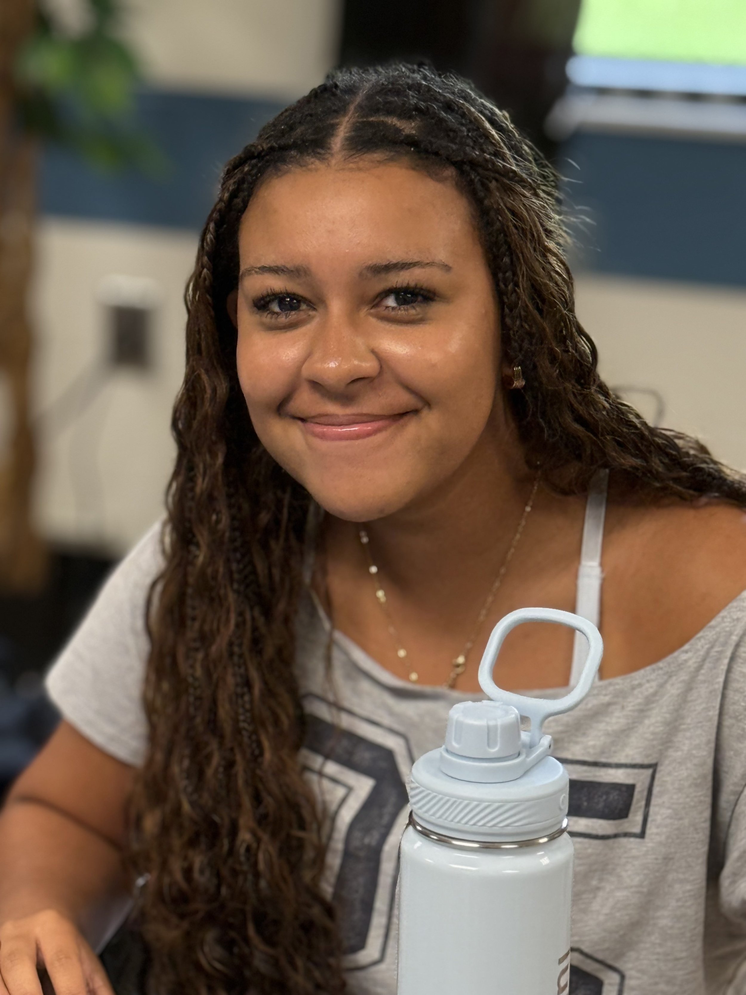 A smiling woman with long, curly hair sitting at a table with a water bottle in front of her.