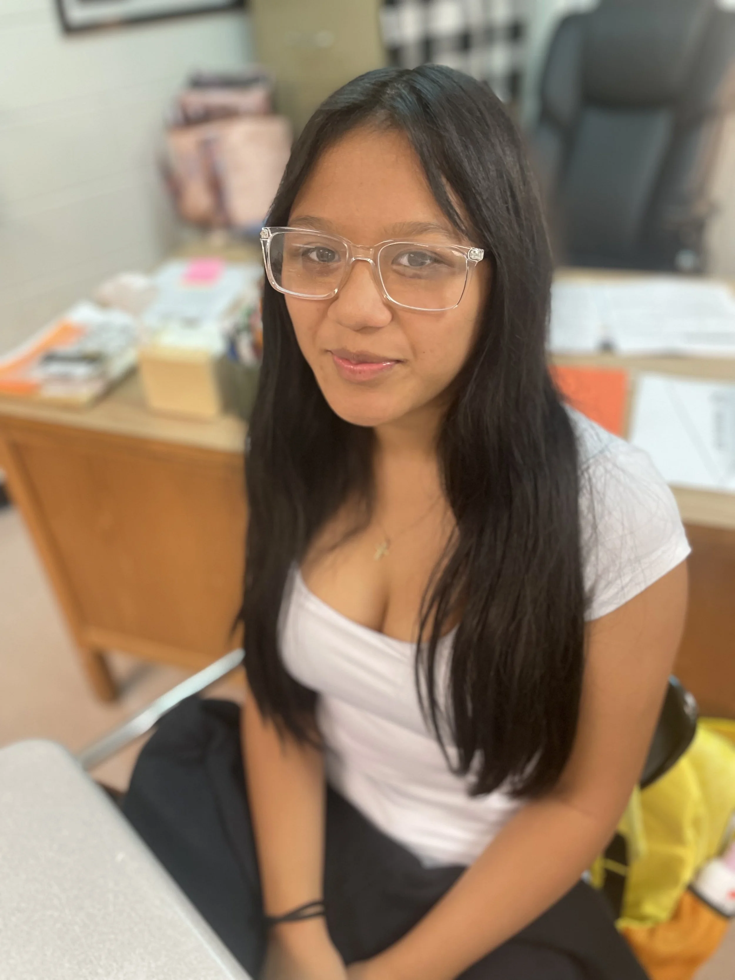 A young woman with long black hair, wearing glasses and a white top, sitting indoors at a desk with various objects and books in the background.