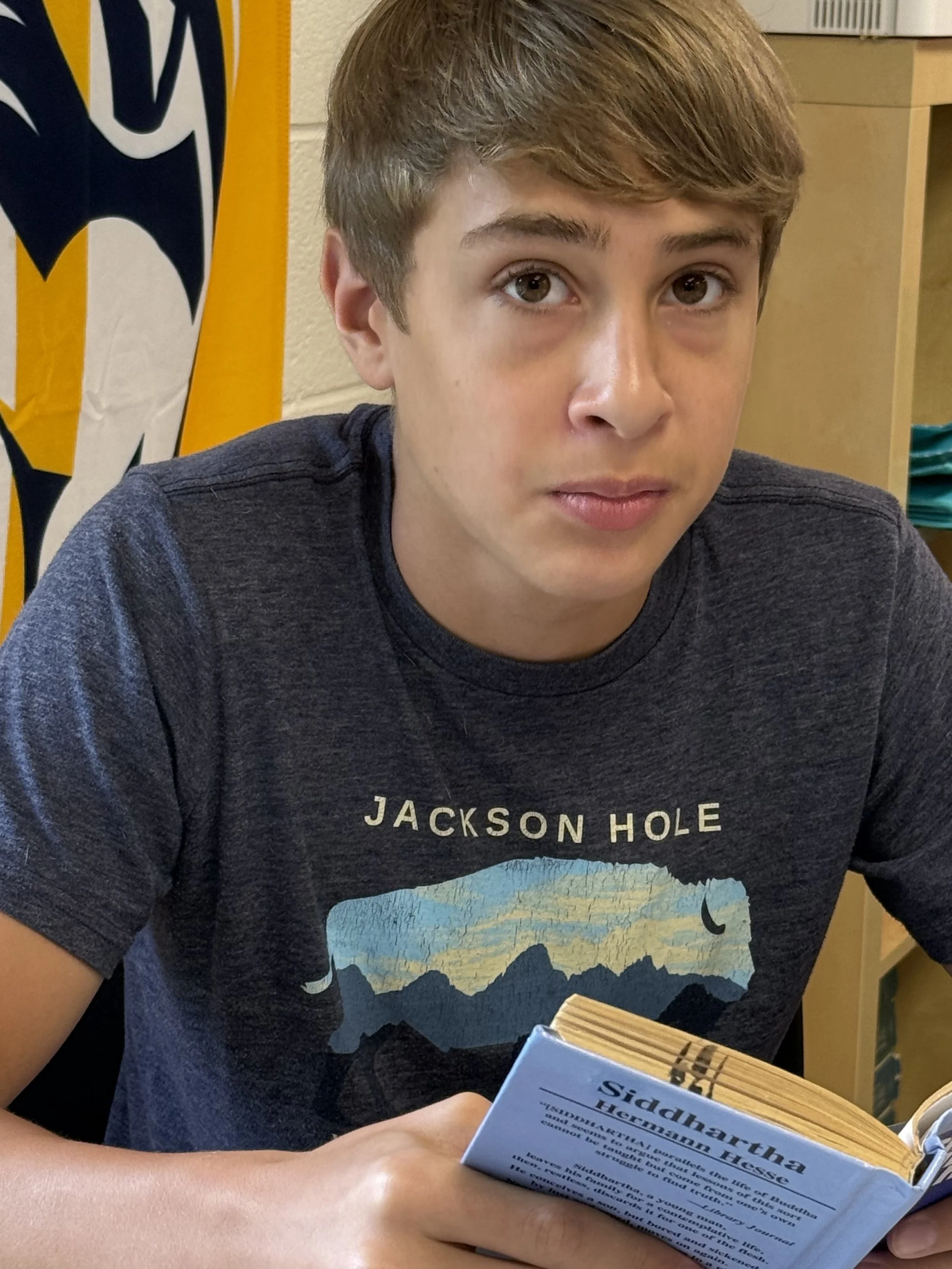 A young boy with brown hair and wearing a blue shirt looks up at the camera in a classroom, sitting at a desk with a green notebook and a pencil in front of him. Other students are visible in the background.