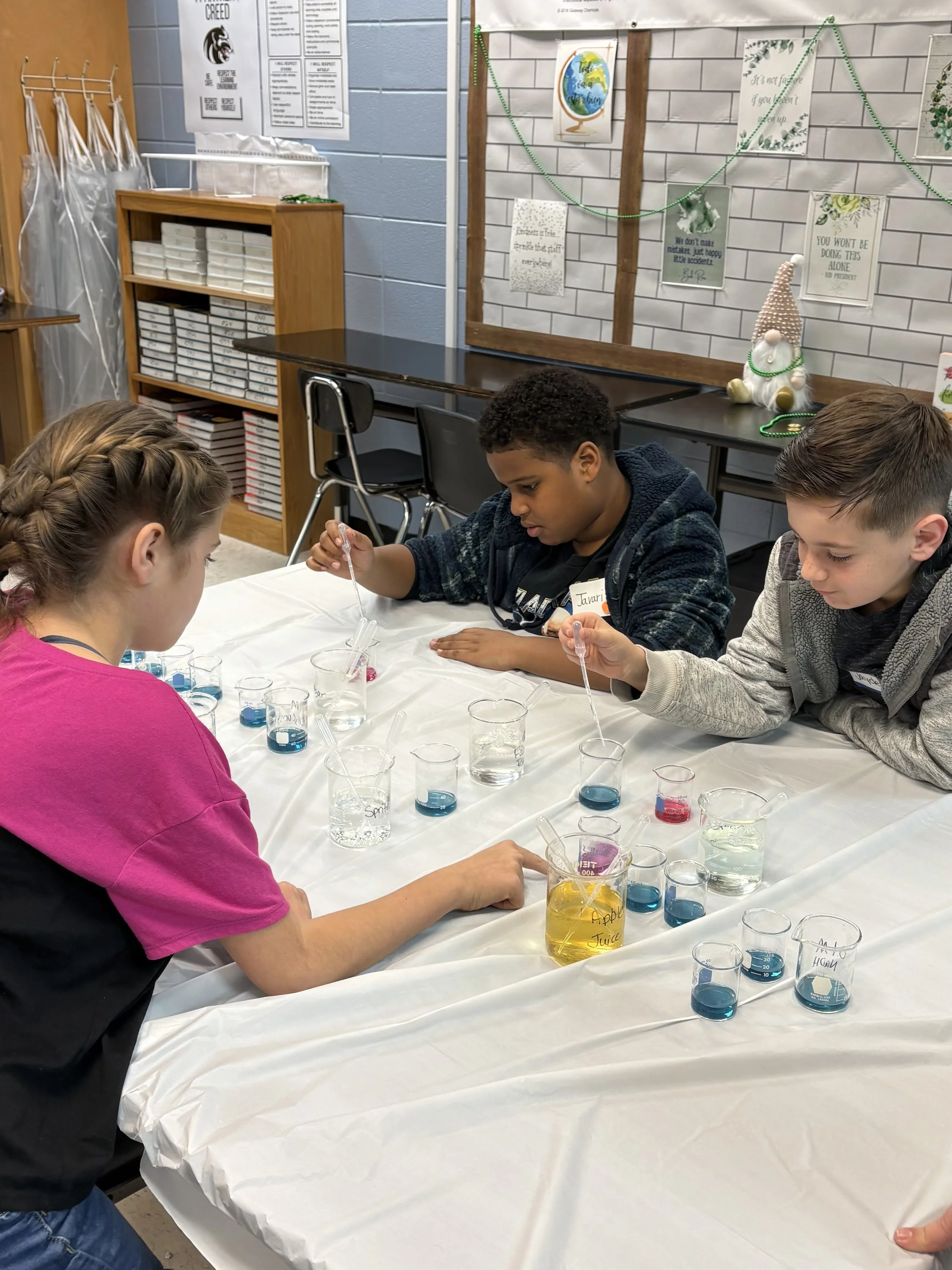 Three children conducting a science experiment with test tubes filled with colorful liquids at a classroom table.