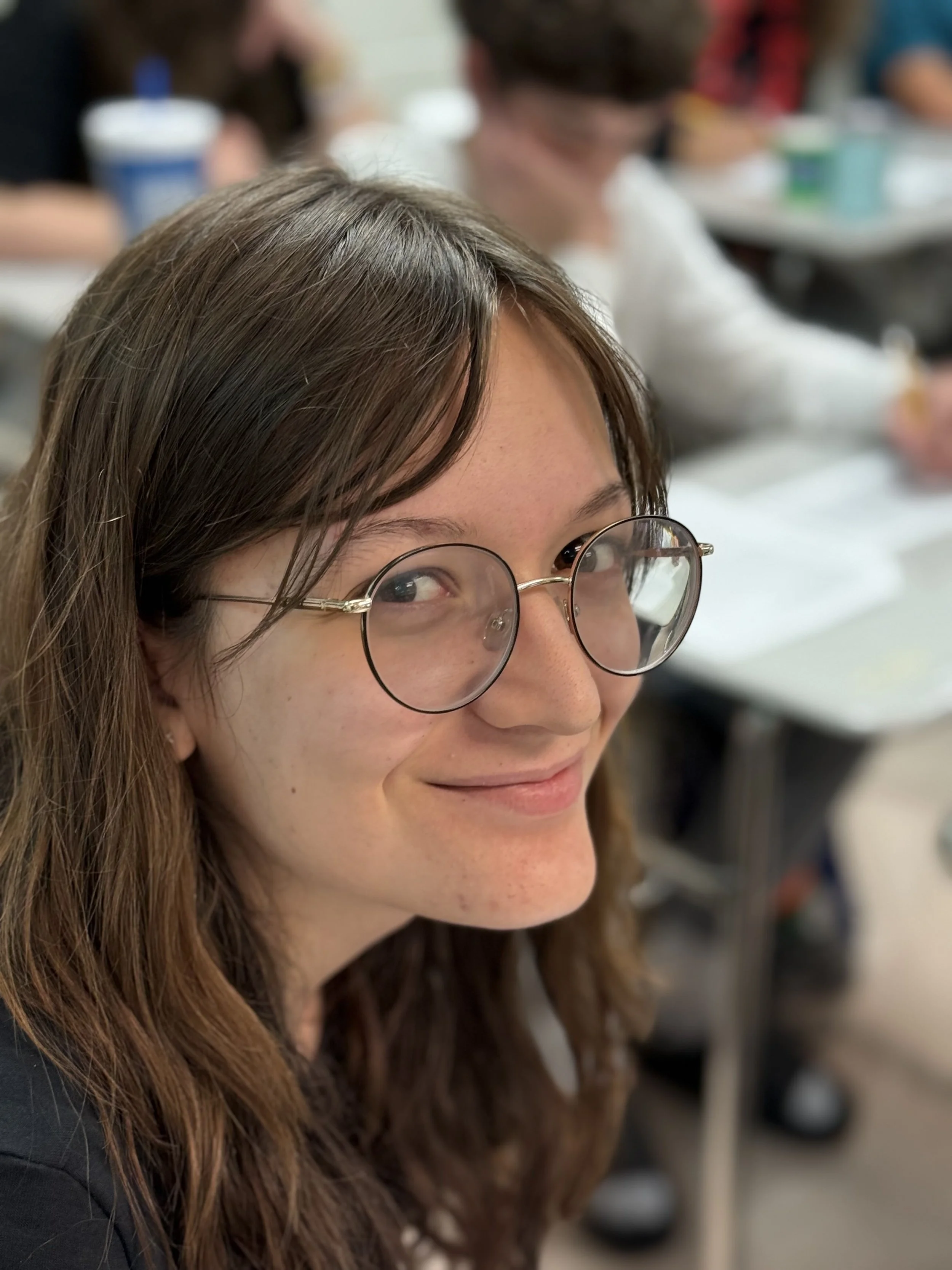 A young woman with brown hair, glasses, and freckles is smiling in a classroom setting.