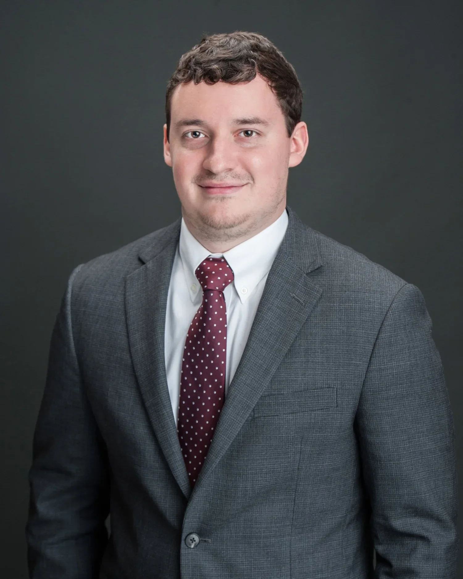 Portrait of a young man in a gray suit, white shirt, maroon polka dot tie, with dark, short, curly hair, smiling softly against a dark gray background.