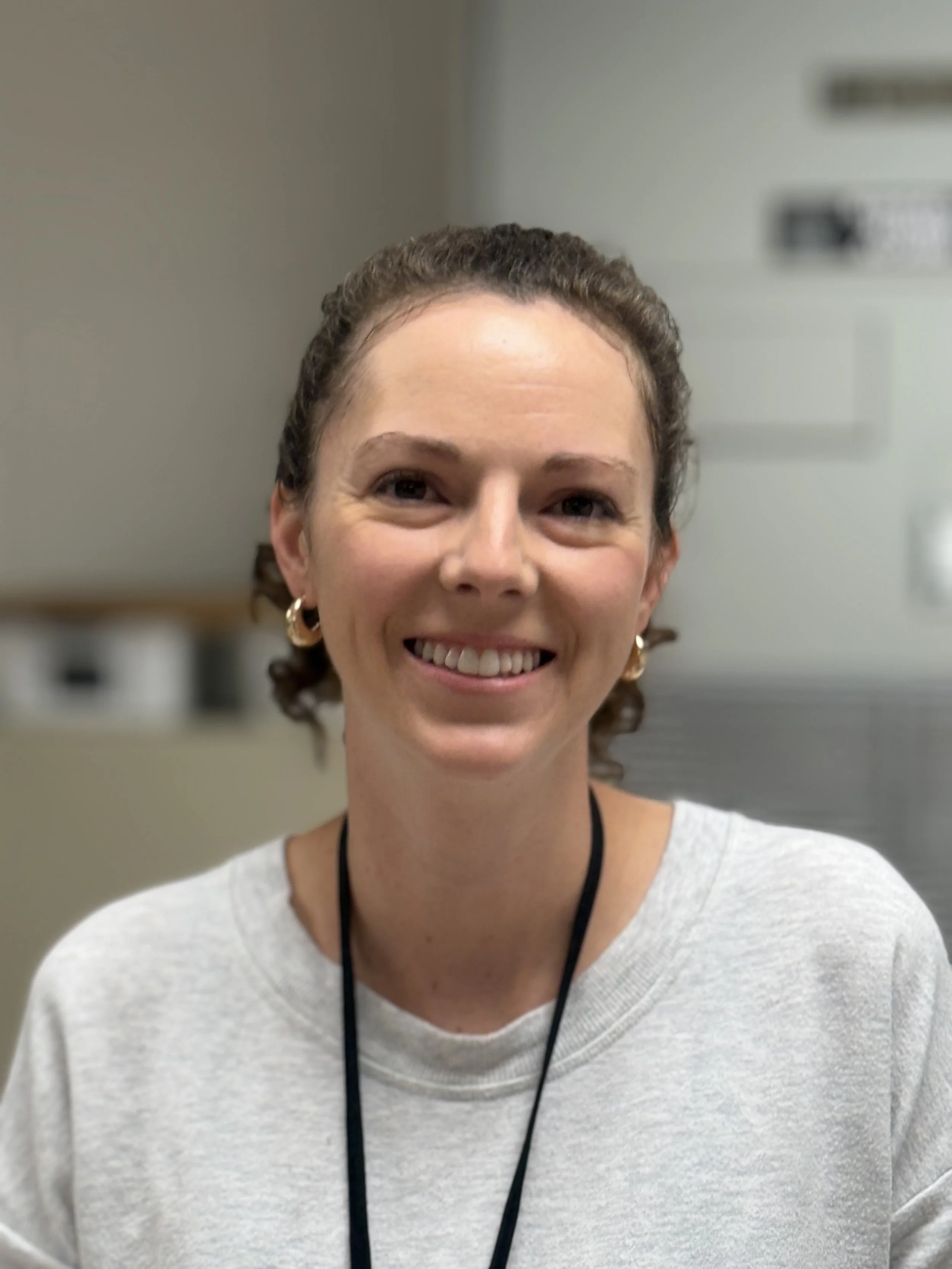 A woman with curly brown hair, gold hoop earrings, and a light grey sweatshirt smiling at the camera in an indoor setting.