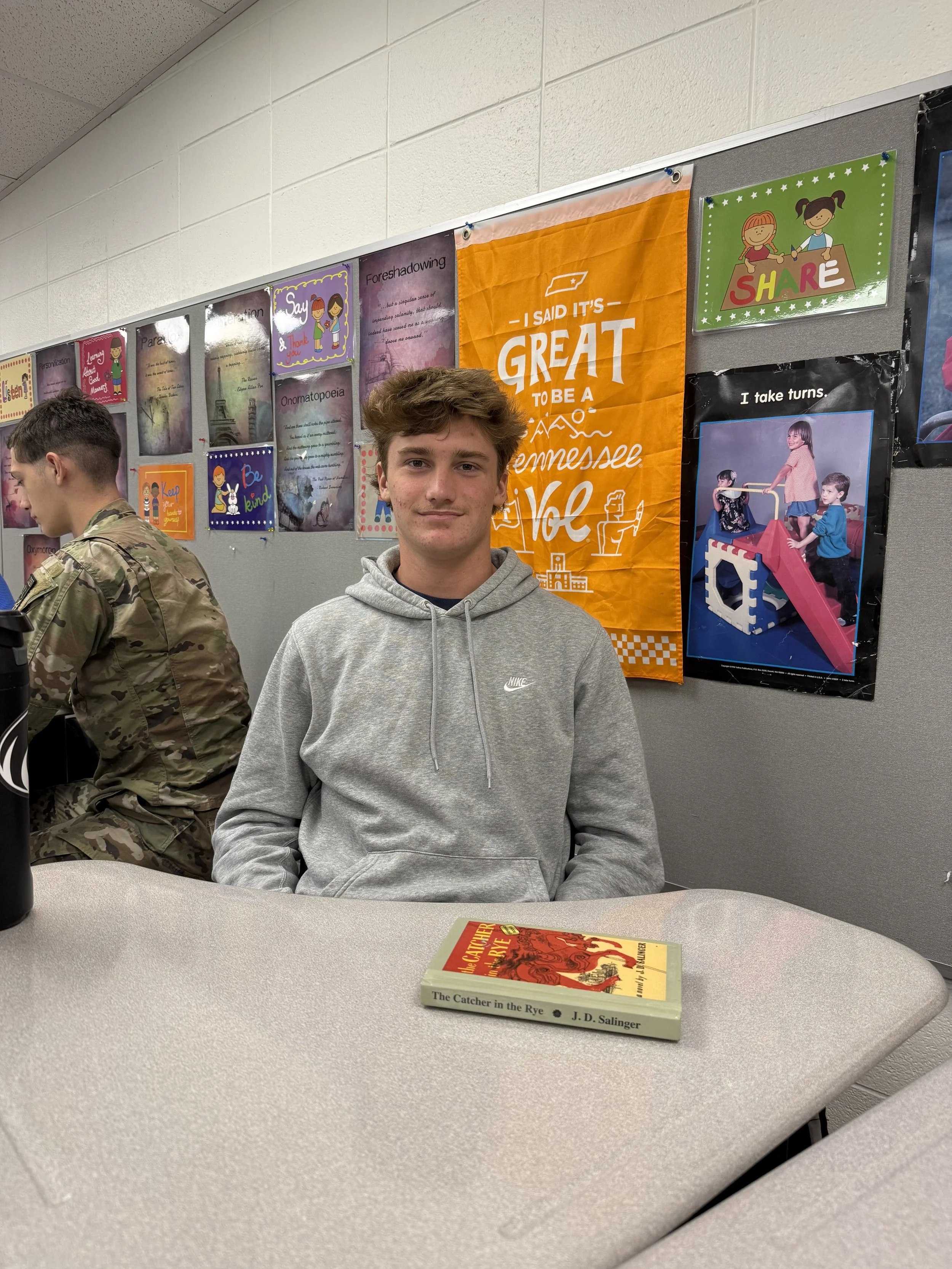 A teenage boy with reddish-brown hair sitting at a desk in a classroom, looking at the camera, wearing a black Nike windbreaker, a smartwatch, and a bracelet, with other students in the background.