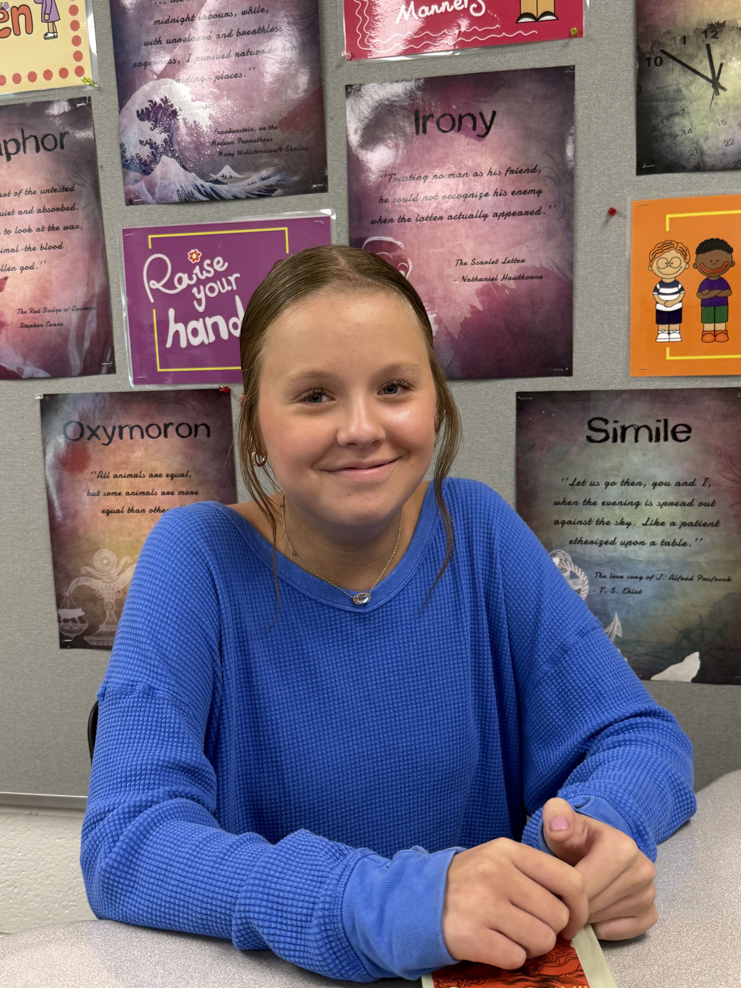 A smiling girl with braces sitting at a table in a classroom or office, with a Chromebook in front of her and a beige filing cabinet behind her.