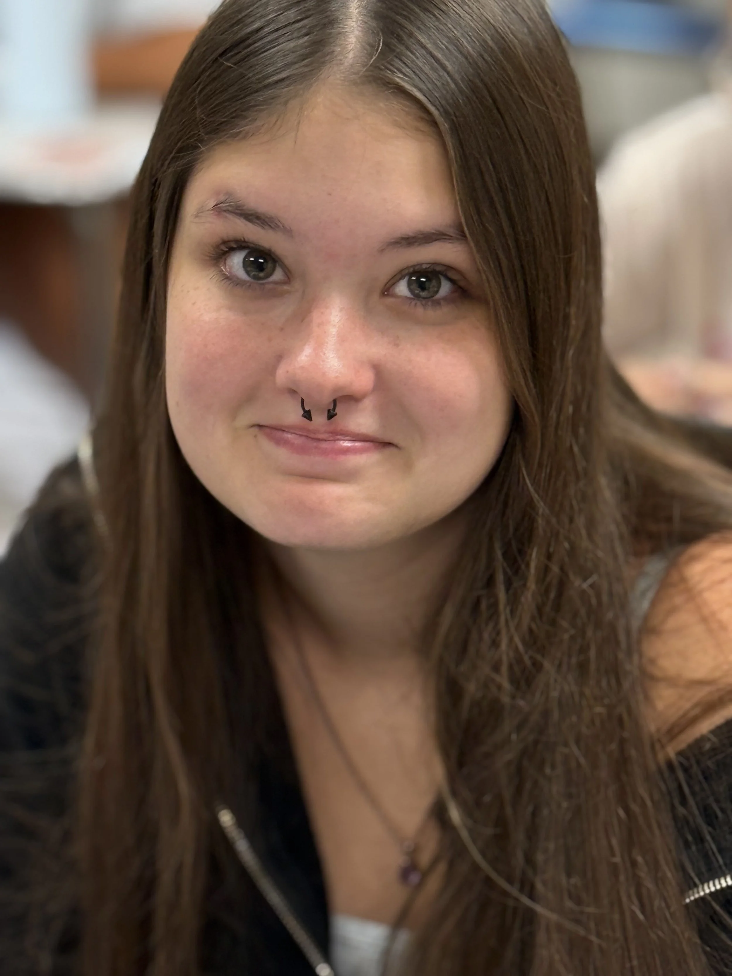 Close-up of a young woman with long brown hair, wearing a septum piercing, smiling slightly, in an indoor setting.