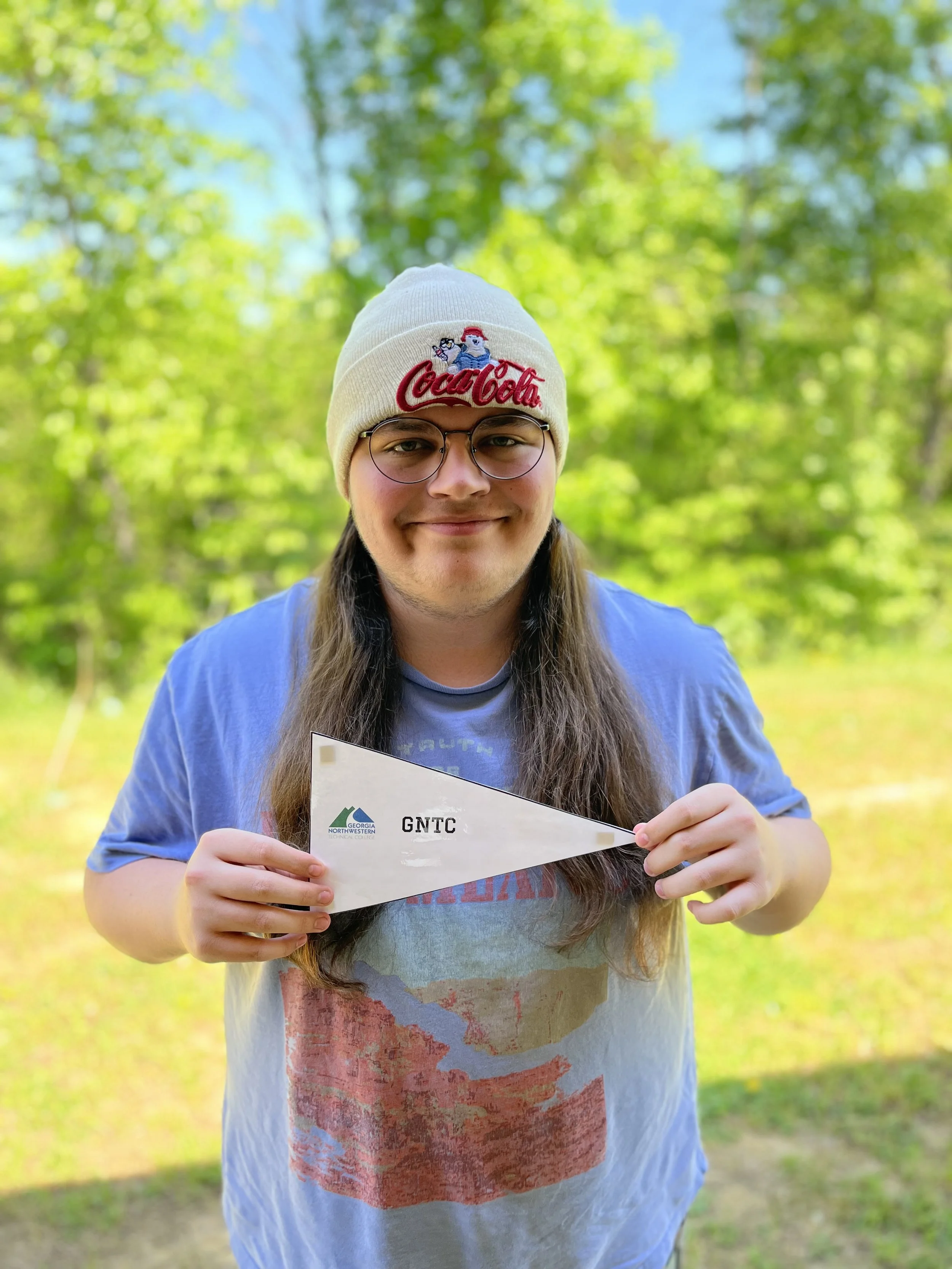 Close-up of a young person with long brown hair, wearing glasses, smiling, with a black t-shirt featuring a white mountain graphic.