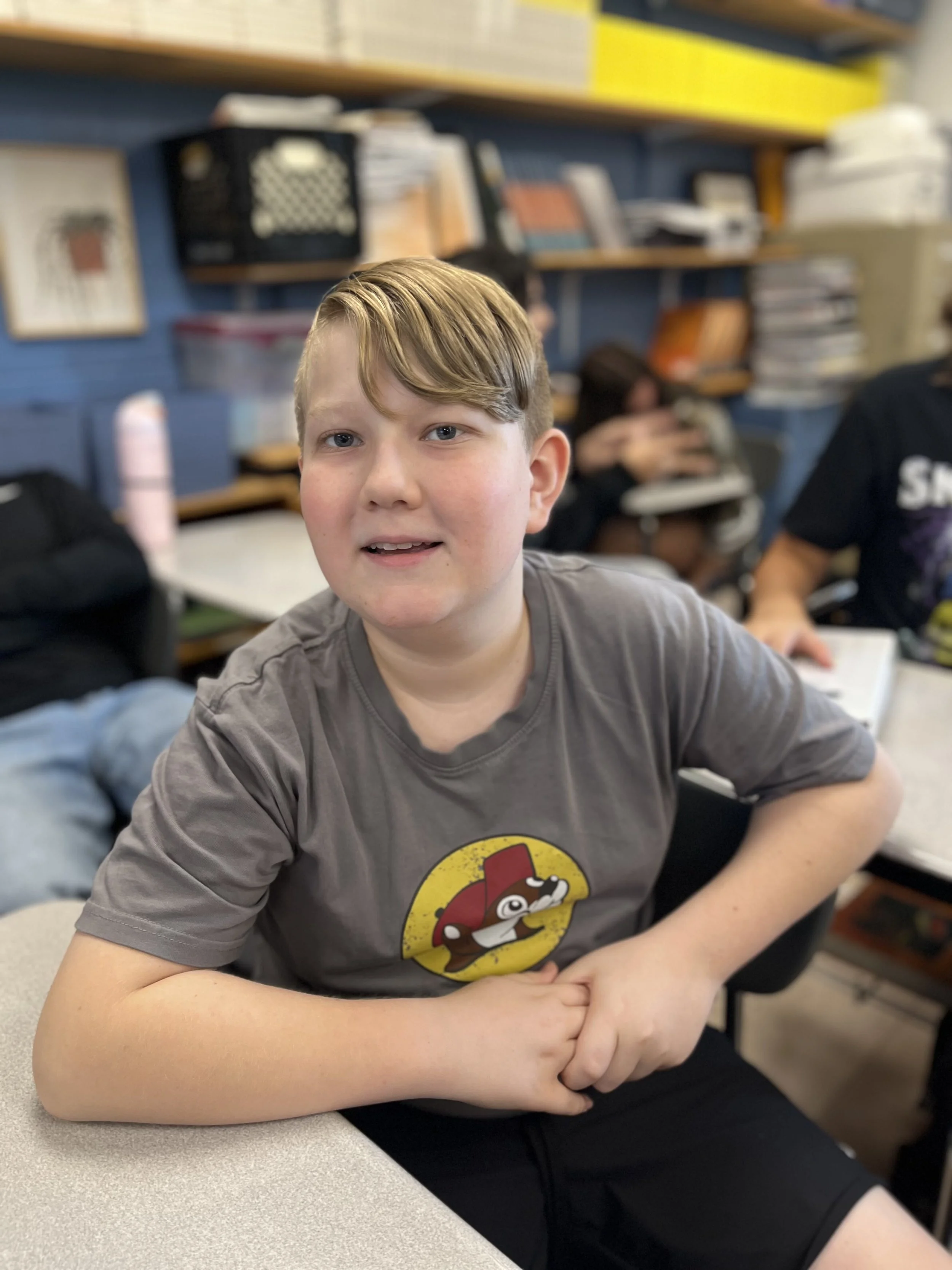 A teenage boy with brown hair and fair skin sitting at a desk in front of a whiteboard with red writing, wearing a blue t-shirt, looking directly at the camera with a serious expression.