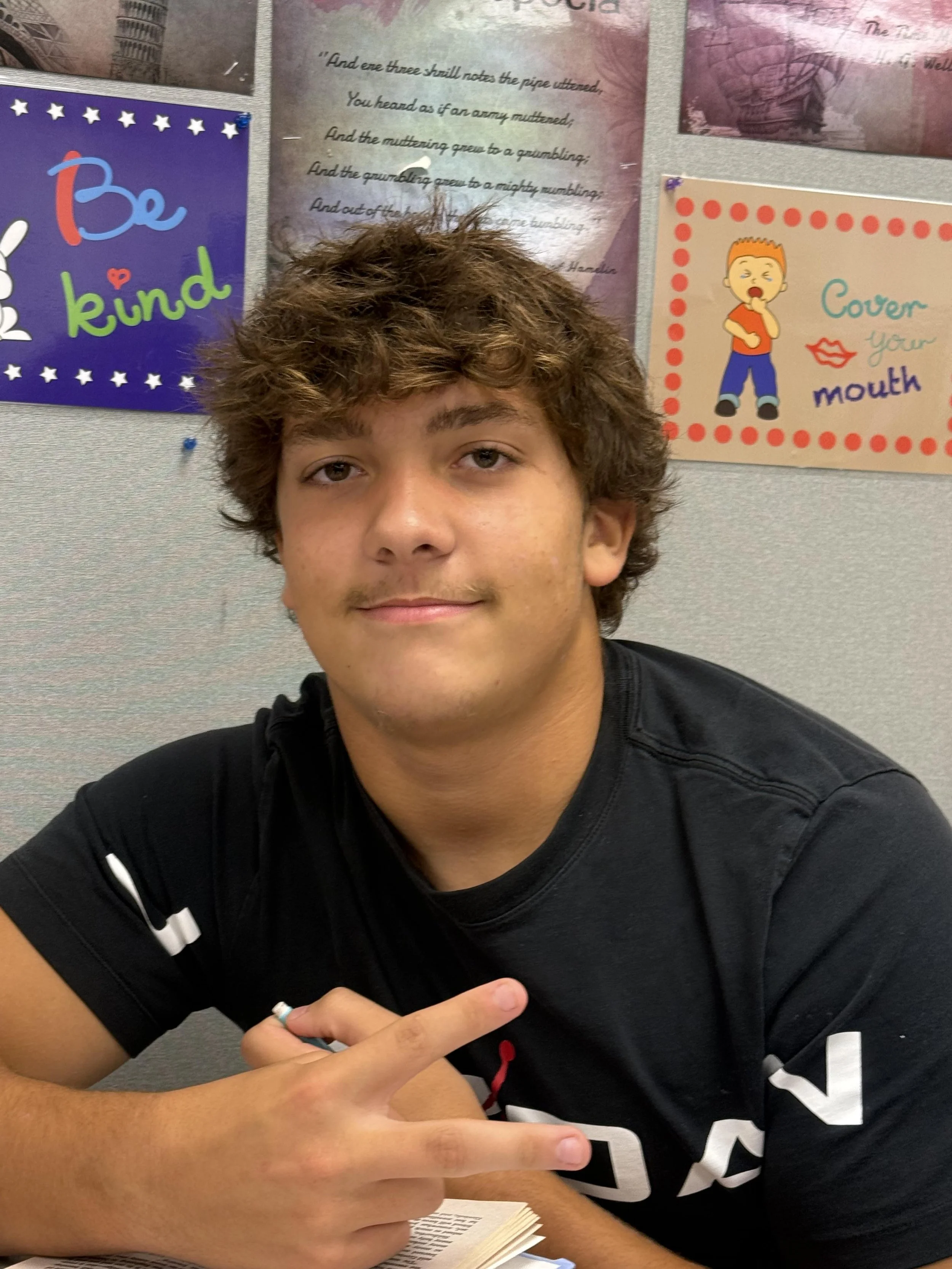 A young man with dark curly hair and light skin touches his chin with his hand, smirking slightly while looking at the camera.