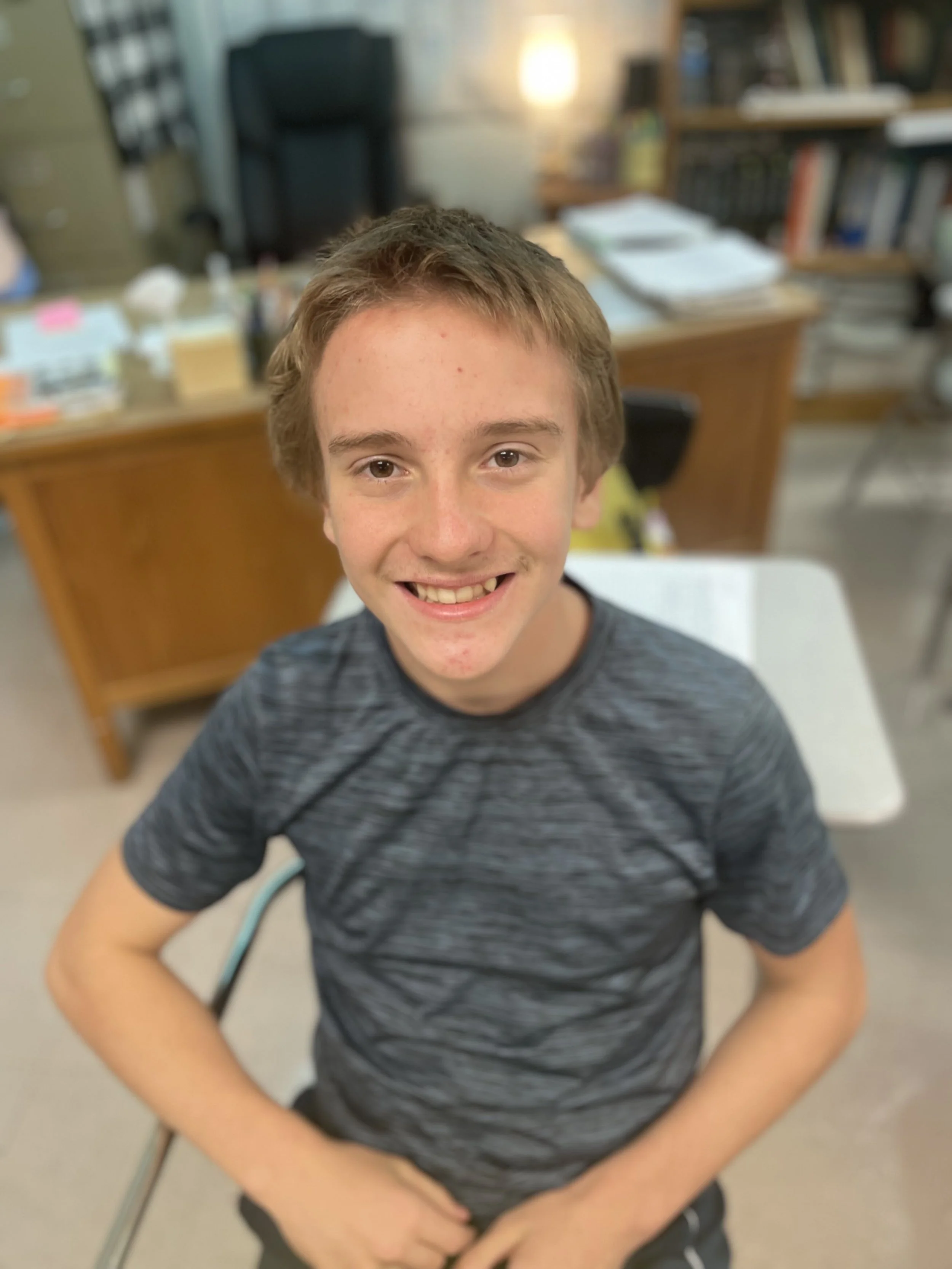 A smiling young boy with short light brown hair, wearing a dark gray T-shirt, sitting in a classroom or office environment with shelves, books, and a desk in the background.