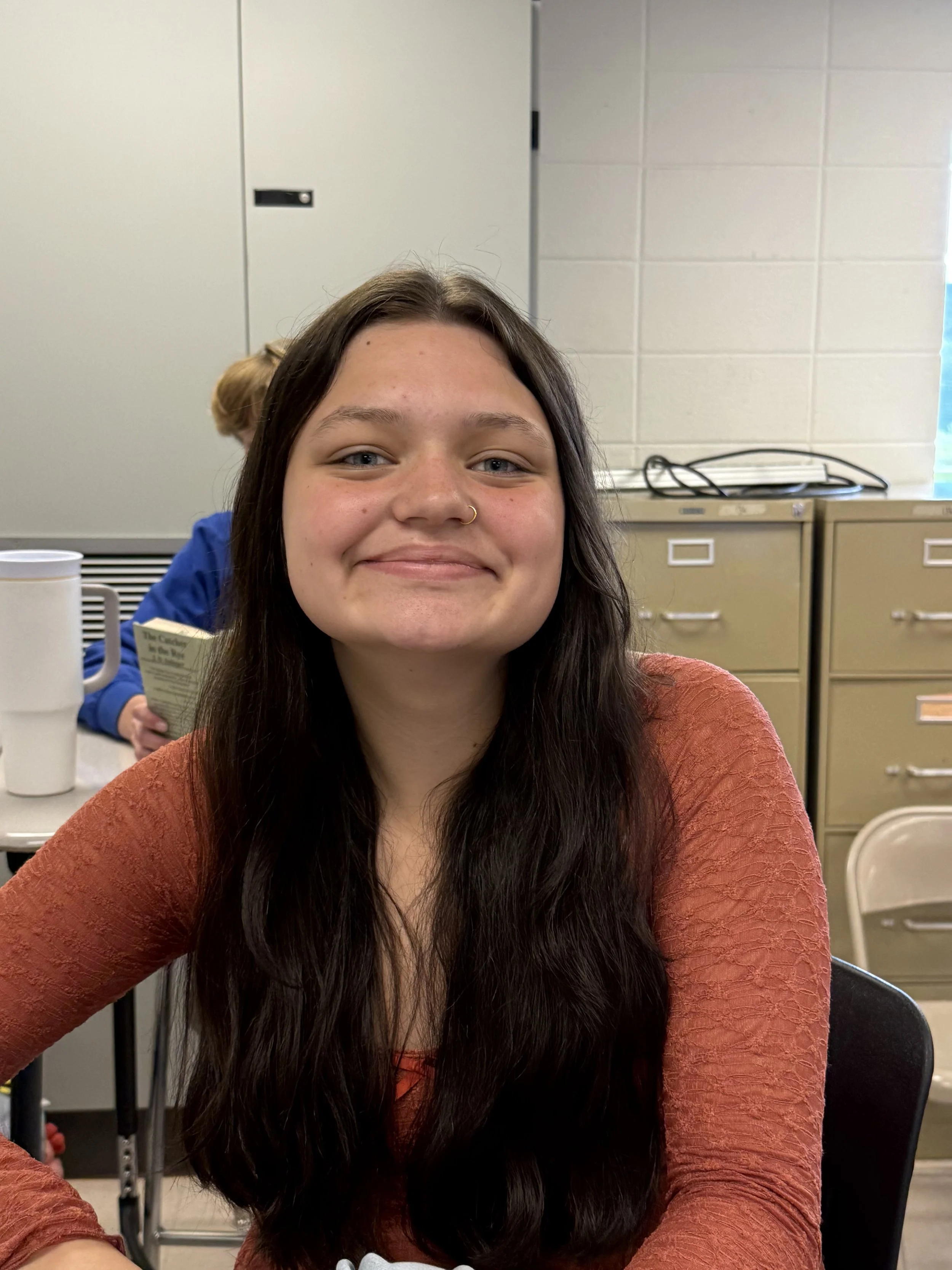 A young girl with long brown hair, wearing a blue Adidas hoodie, smiling at the camera in a classroom setting with other students in the background.