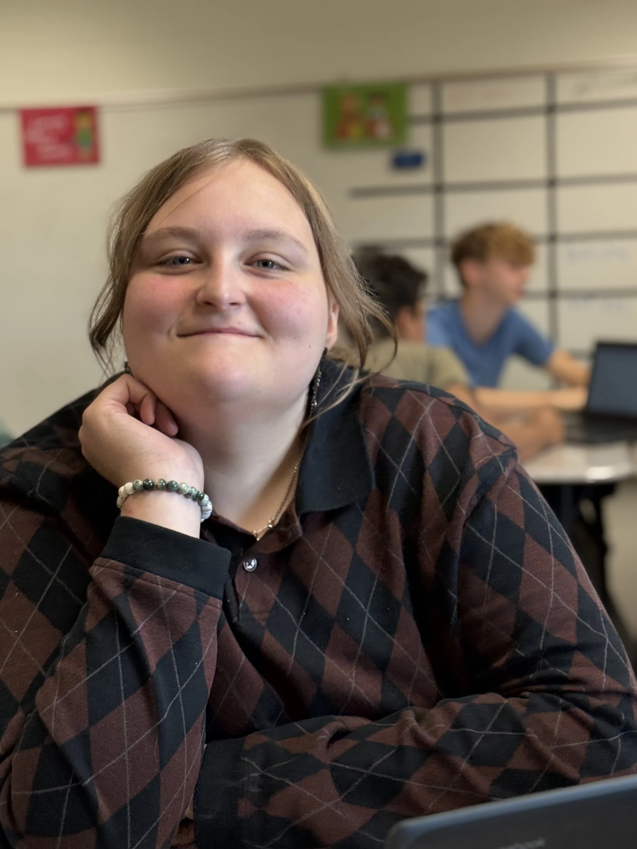 A smiling girl with light brown hair, wearing a black and red plaid shirt, sitting at a desk with a hand resting on her chin, in classroom setting with two boys in the background working on laptops.