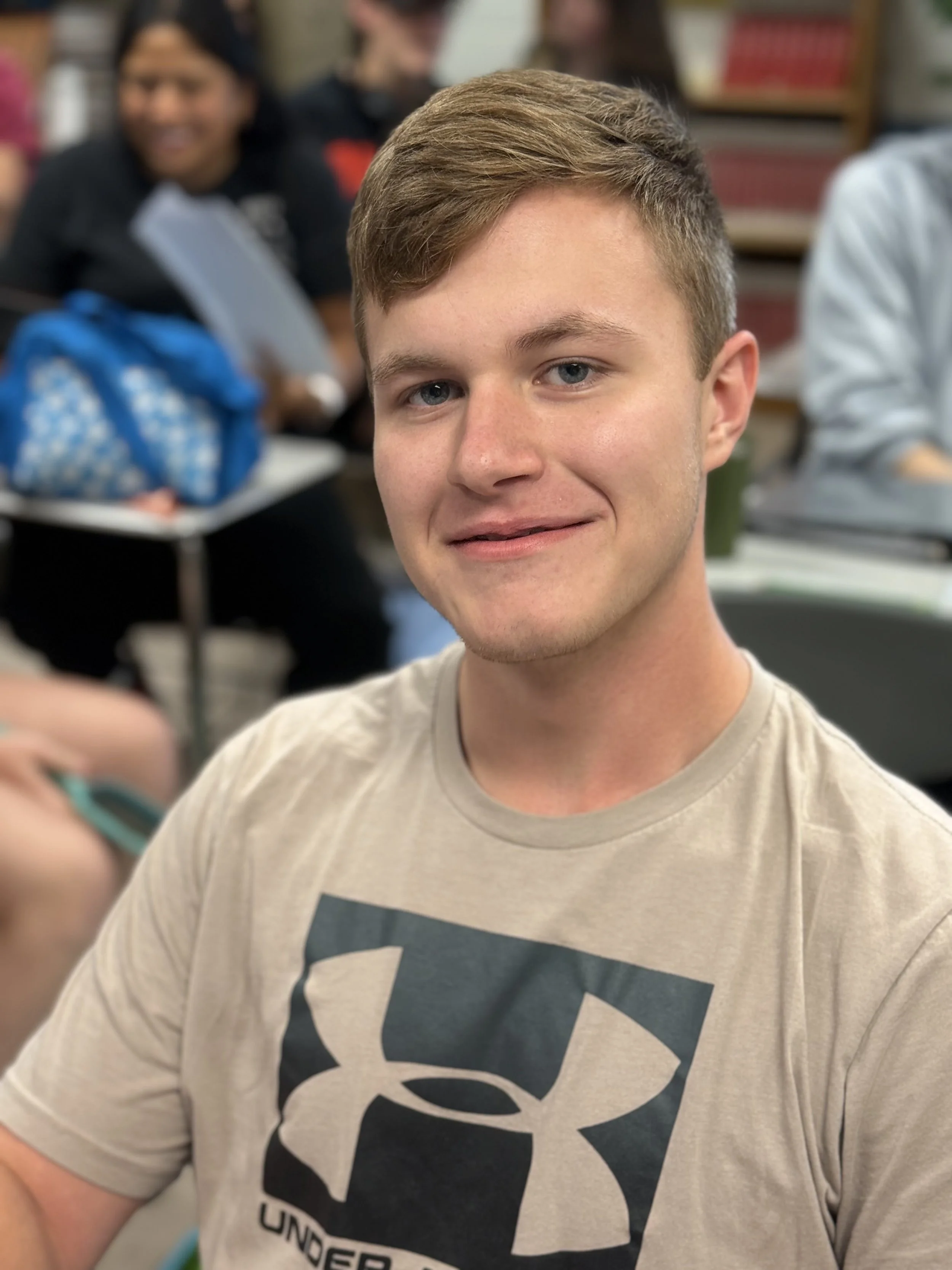 Young man with short light brown hair smiling in a classroom or library setting with blurred background showing other students, bookshelves, and a blue backpack.