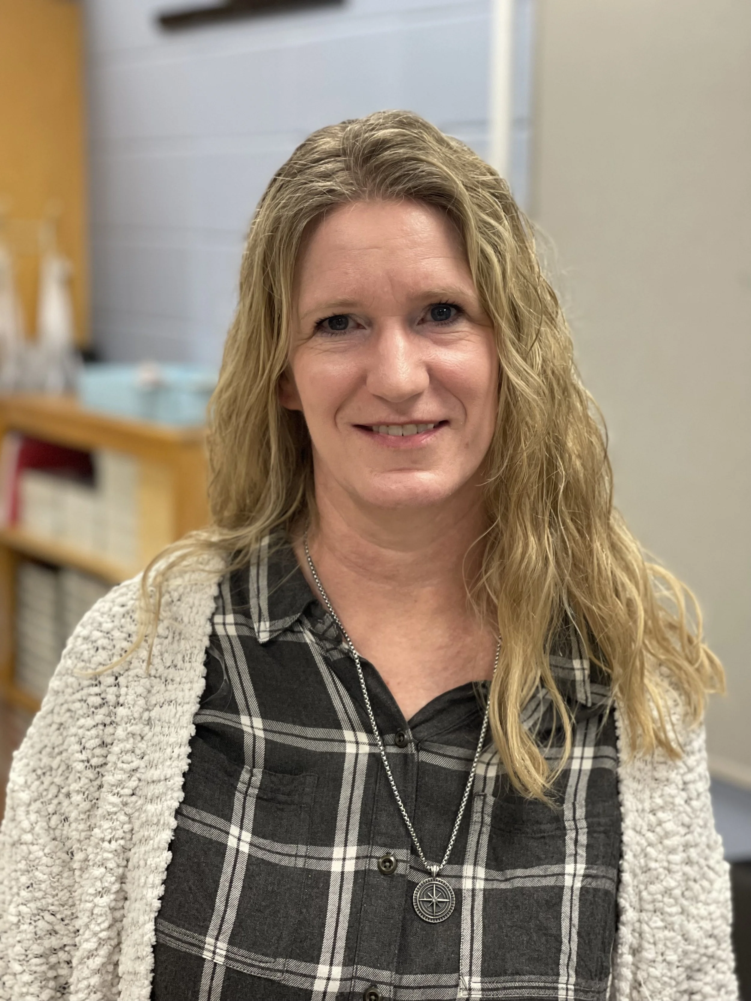 A woman with wavy blonde hair smiling, wearing a black and white plaid shirt with a necklace, in an indoor setting with shelves and boxes in the background.
