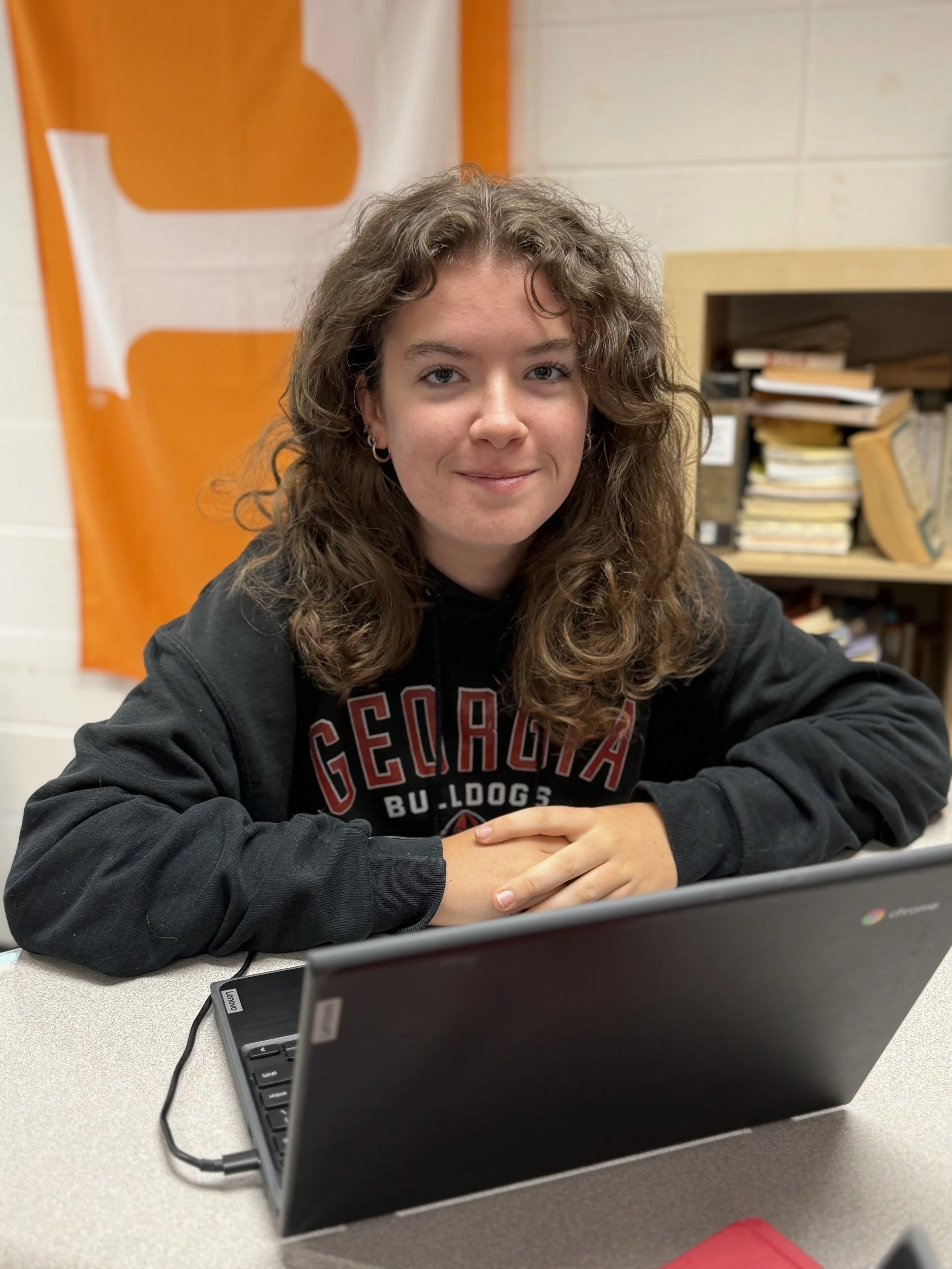 A young woman with curly brown hair and hoop earrings sitting at a desk in a classroom, looking at the camera with a slight smile, in front of a Chromebook laptop, wearing a black sweatshirt with 'Georgia Bulldogs' written on it.