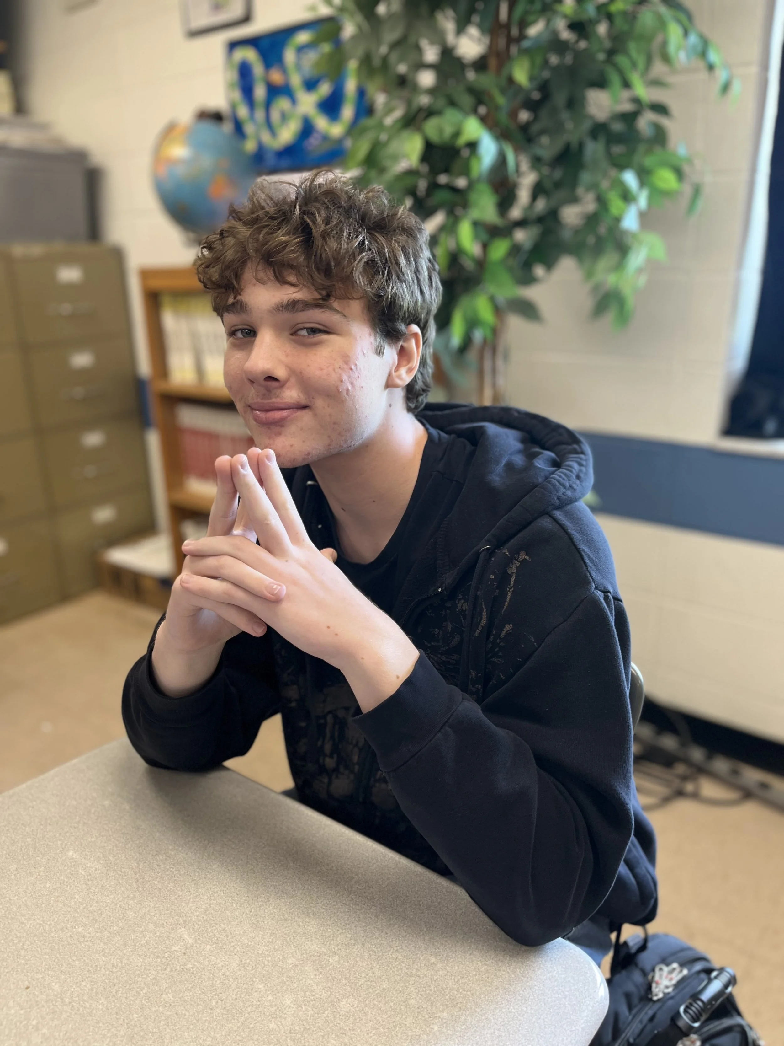 A young man with curly brown hair, wearing a black hoodie, sitting at a desk in front of a laptop, smiling slightly while looking at the camera.
