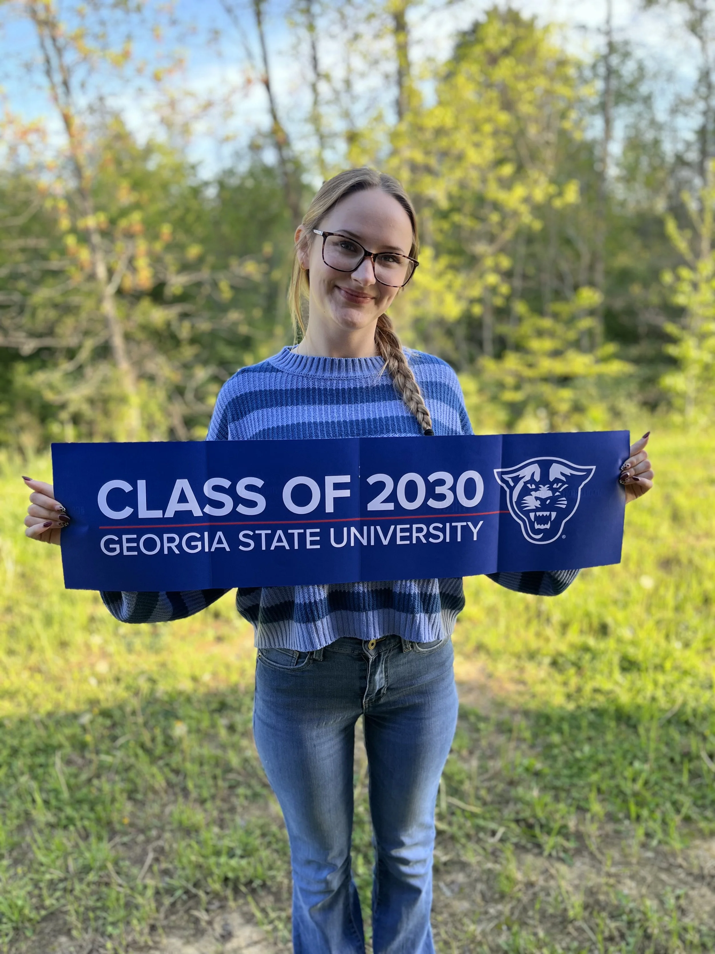 A young woman with long blonde hair, wearing glasses and a black top, smiling at the camera in a classroom or lecture hall setting.