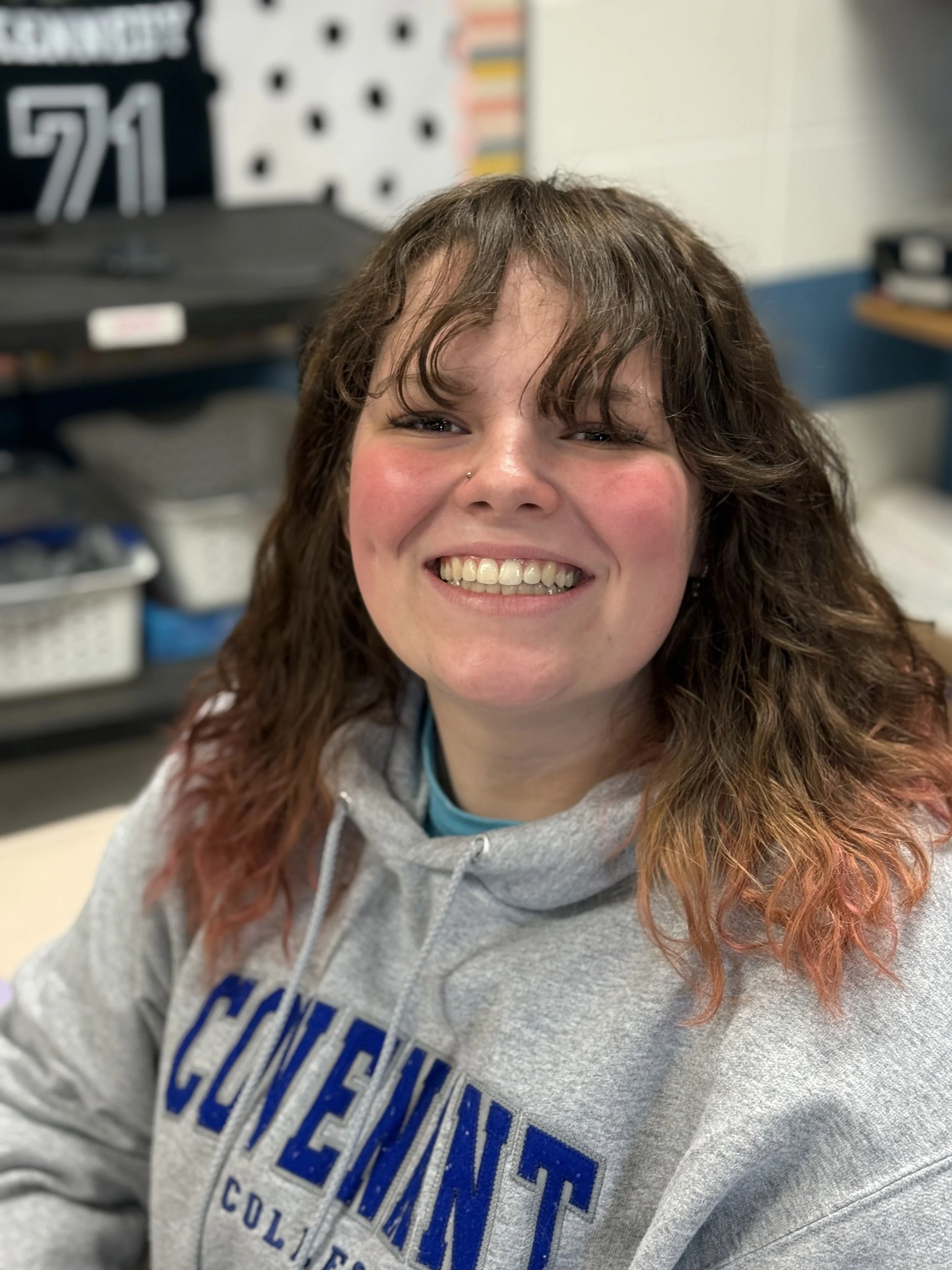 Smiling woman with curly hair wearing a college hoodie in a classroom.
