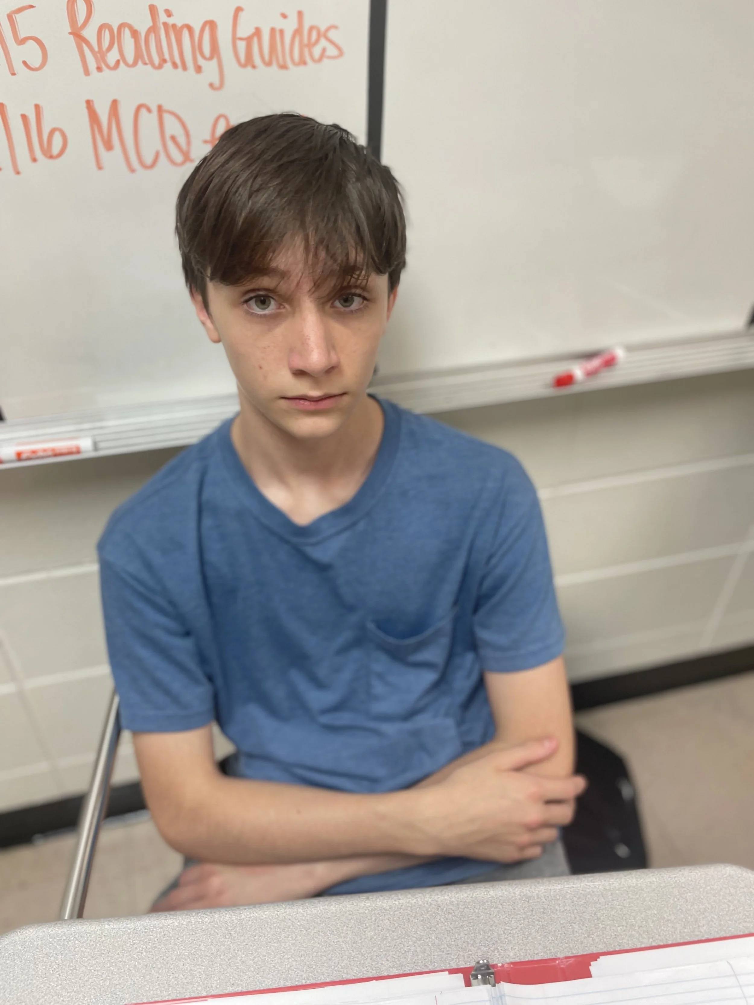 A teenage boy with brown hair and fair skin sitting at a desk in front of a whiteboard with red writing, wearing a blue t-shirt, looking directly at the camera with a serious expression.