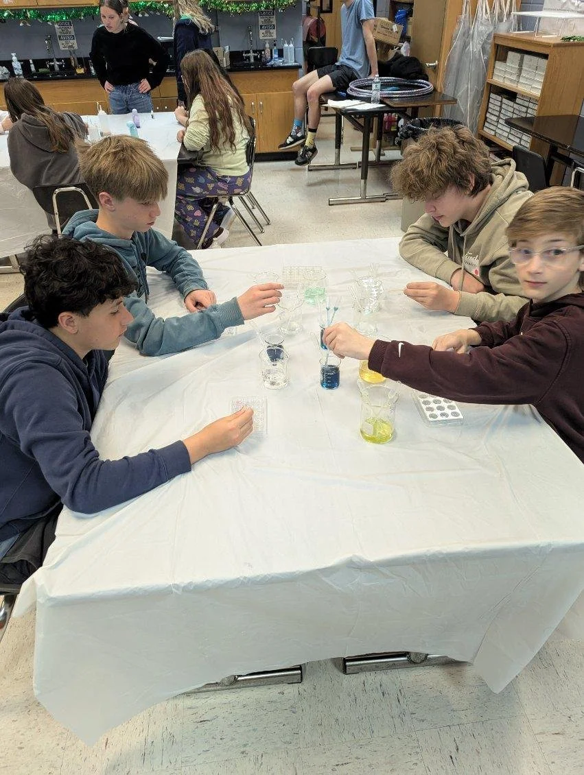 A group of five boys sitting around a table in a classroom or science lab, conducting a science experiment with colored liquids in glass beakers and test tubes. There is a girl and a woman standing in the background.