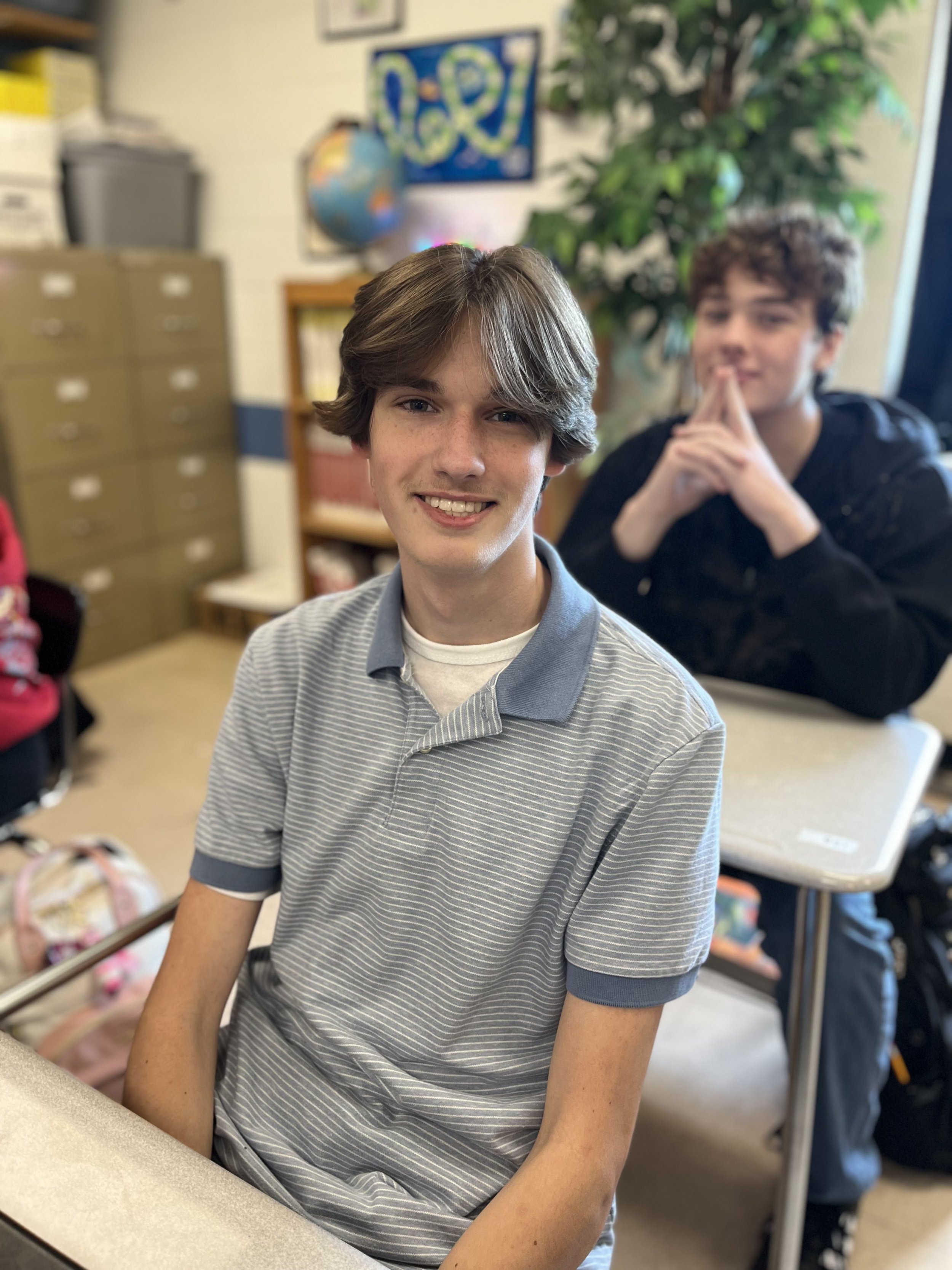 A young person with short brown hair, blue eyes, and fair skin with freckles. They are wearing a gray T-shirt and are in a room with bookshelves and various items in the background.