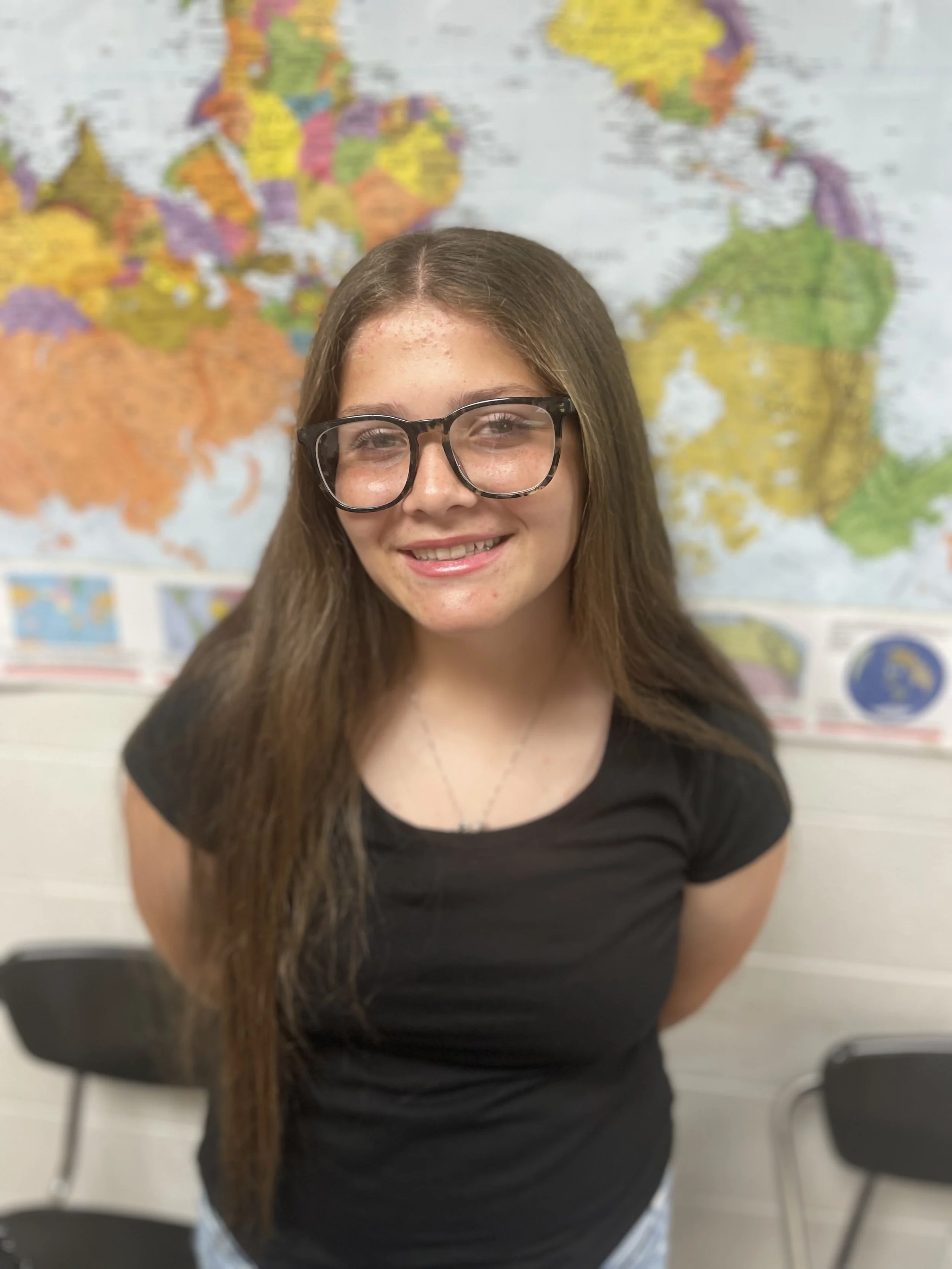 Smiling girl with long brown hair, black glasses, and a black shirt standing in front of a world map in a classroom.