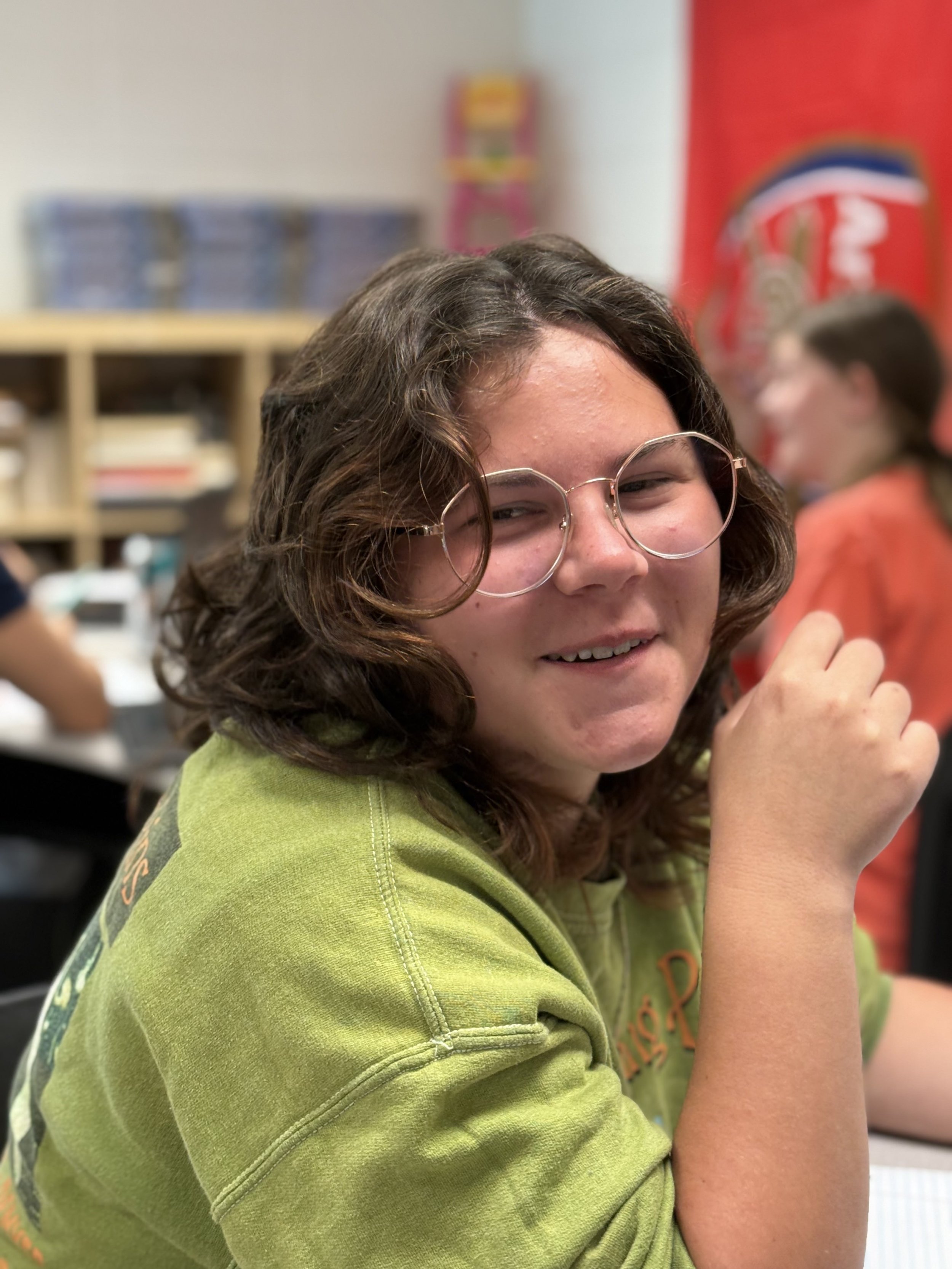 A girl with curly brown hair, glasses, and a green sweatshirt sitting at a table in a classroom, smiling and looking at the camera.