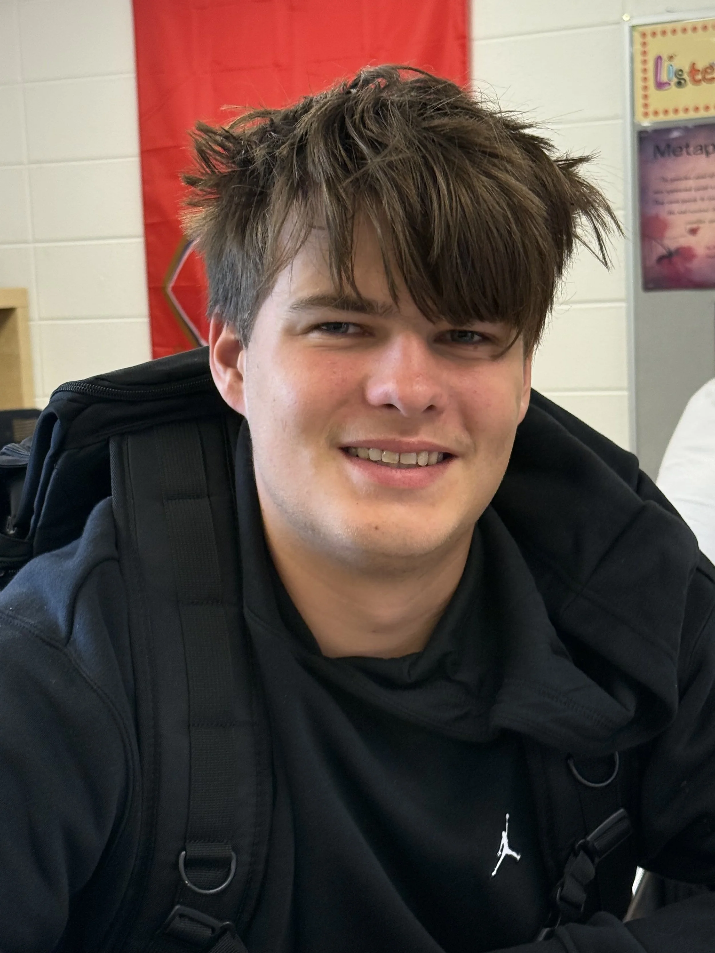 A teenage boy standing in a classroom with other students seated at desks and working on laptops in the background.