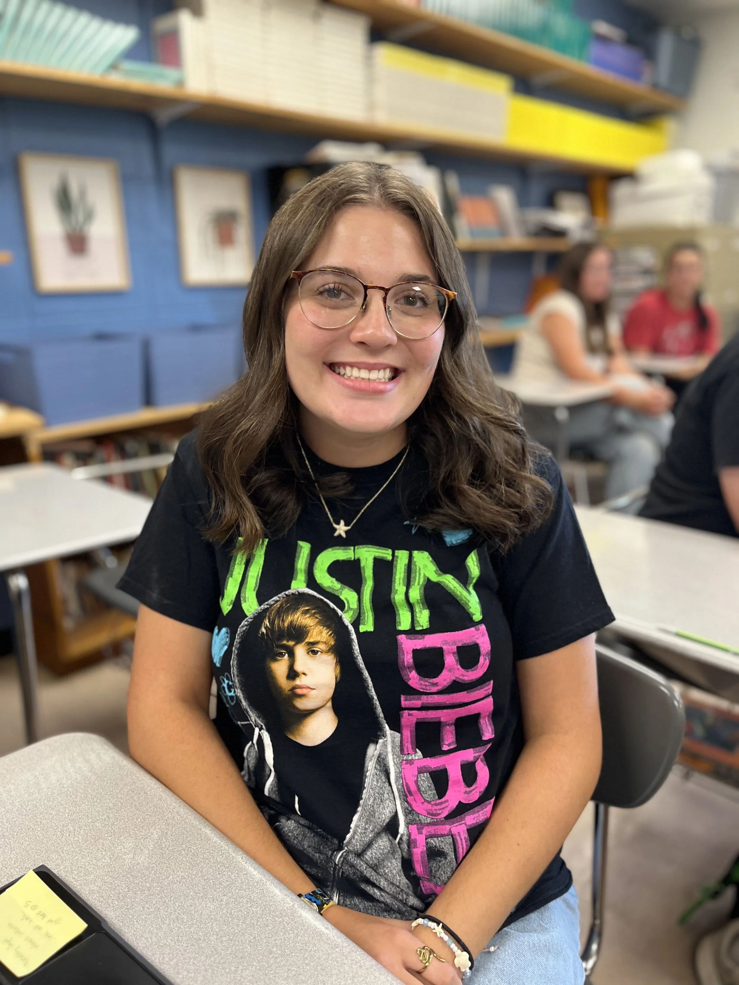 A smiling young woman with glasses and shoulder-length dark brown hair sitting in a classroom, wearing a black 'The Amazing Spider-Man' T-shirt, with others working at desks in the background.