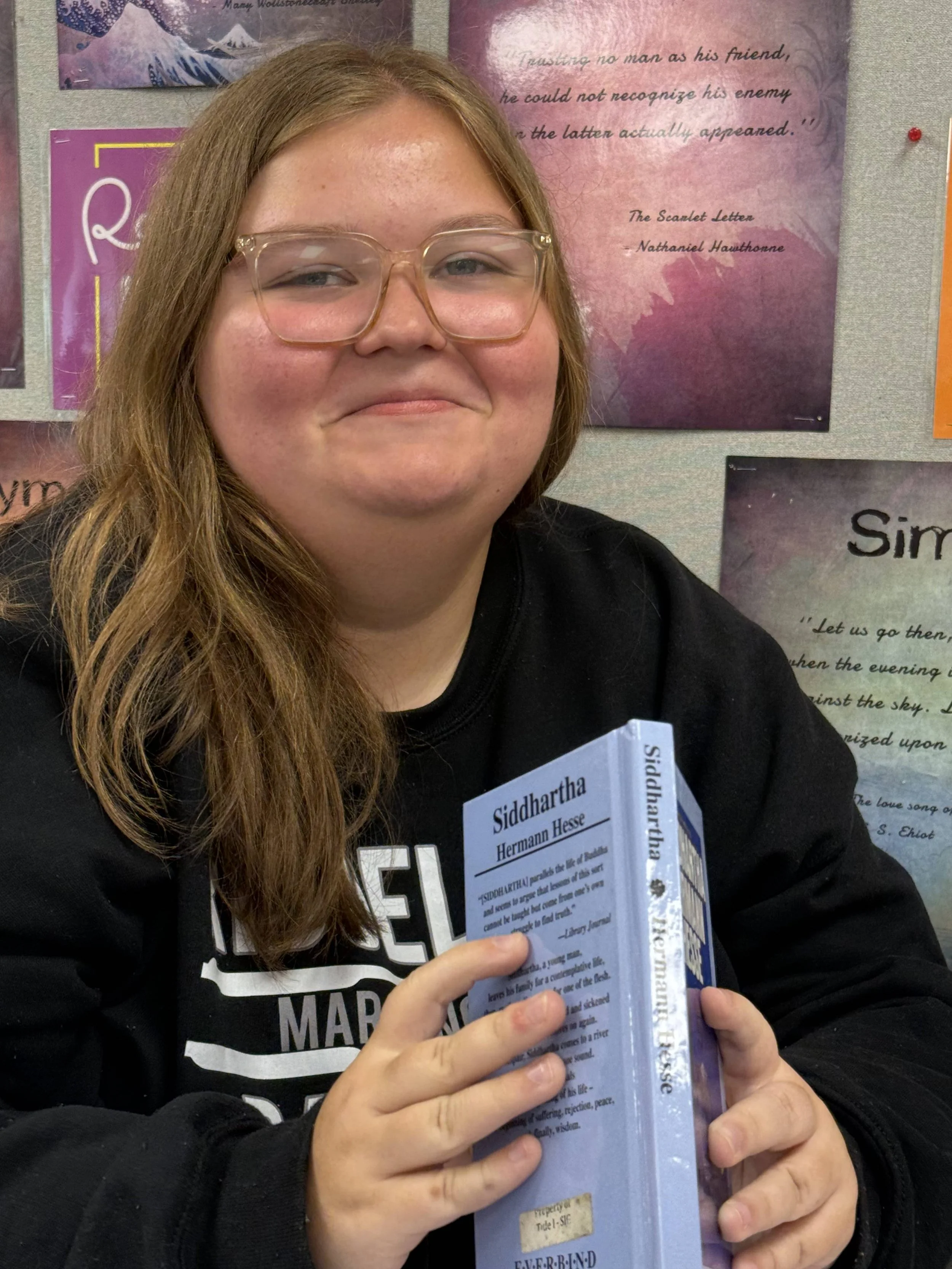 A girl with glasses and brown hair sitting at a desk in a classroom, smiling at the camera with students and teacher in the background.