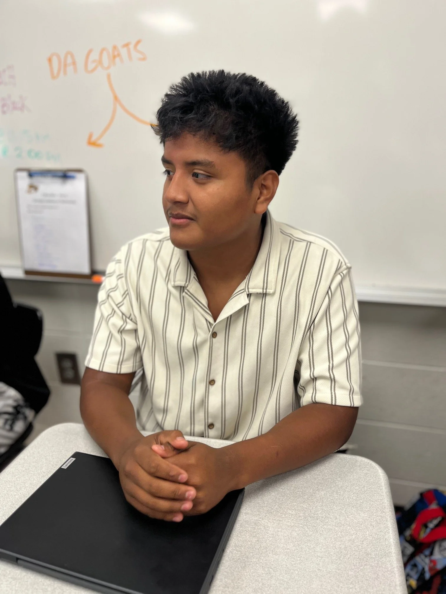 A young man with short dark hair sitting at a desk, looking thoughtfully out a window. He is wearing a gray Aeropostale t-shirt and has a notebook with handwritten notes and a closed Chromebook in front of him.