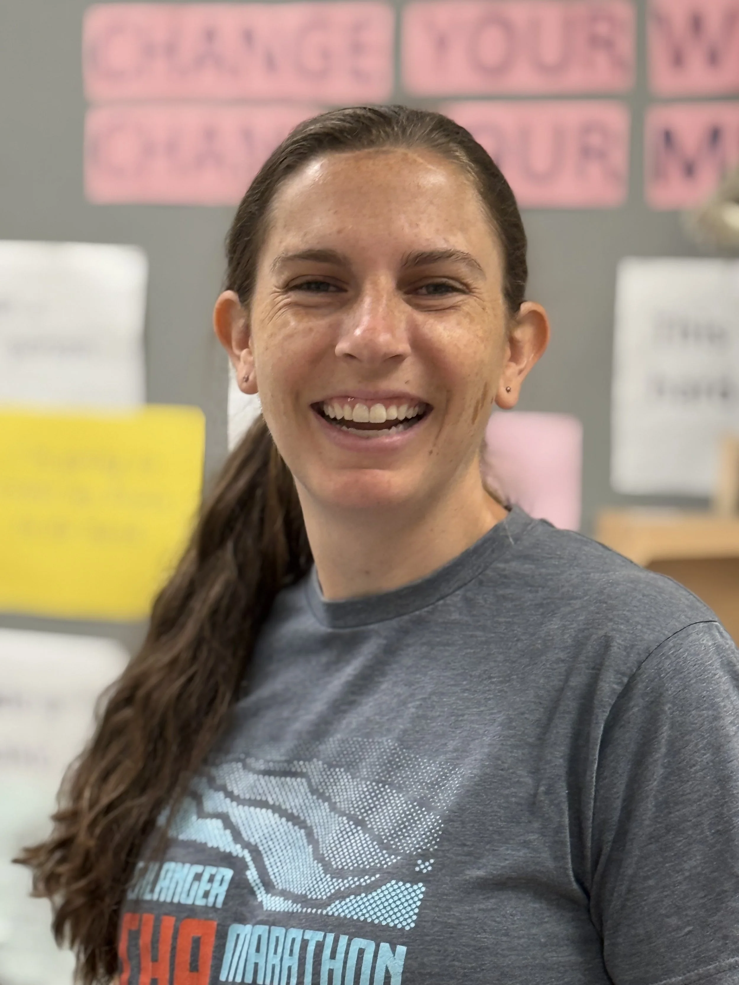 Smiling woman with long brown hair in a ponytail wearing a gray T-shirt with a marathon logo, standing in a room with motivational messages on a wall behind her.