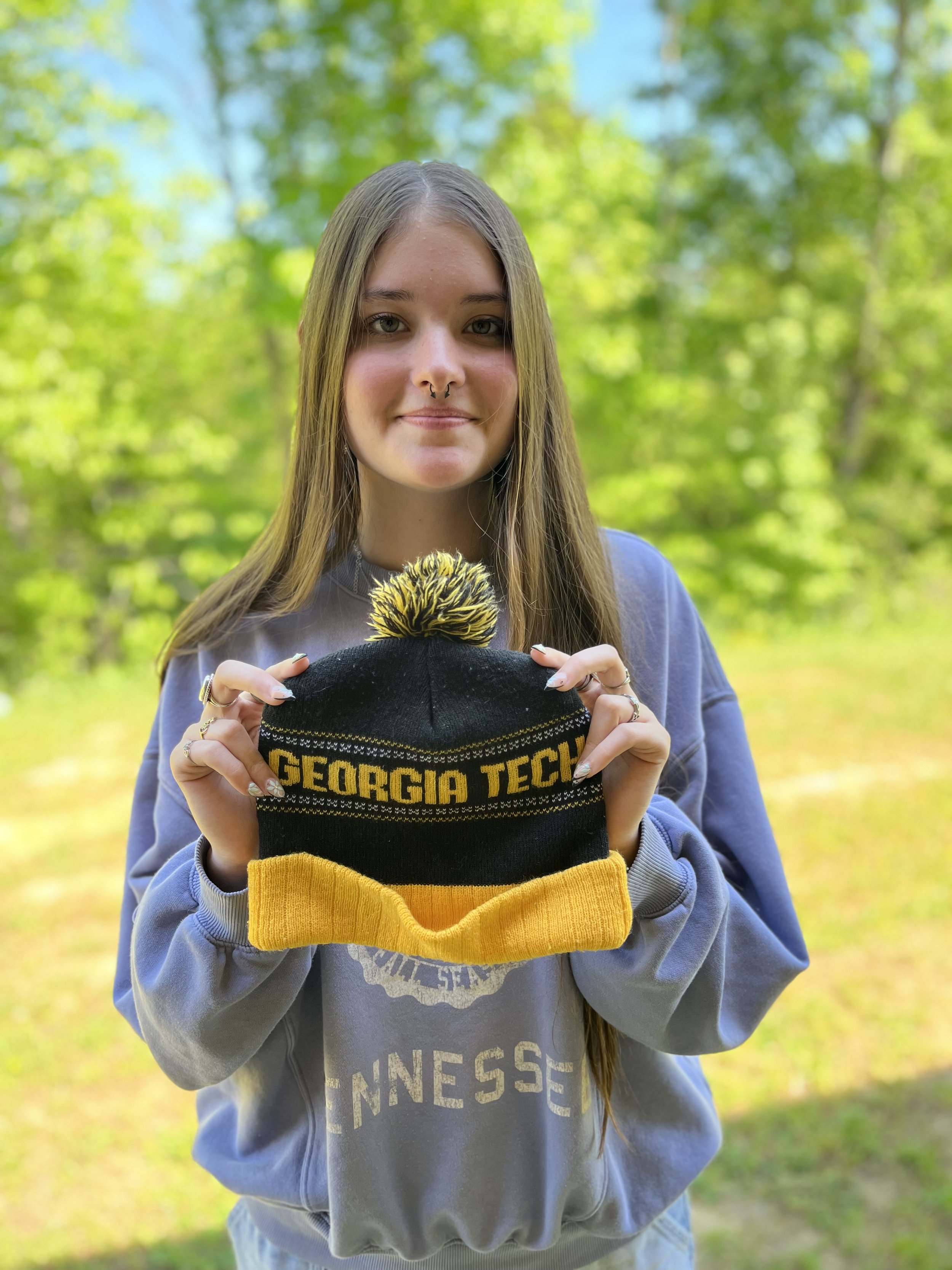 Close-up of a young woman with long brown hair, wearing a septum piercing, smiling slightly, in an indoor setting.