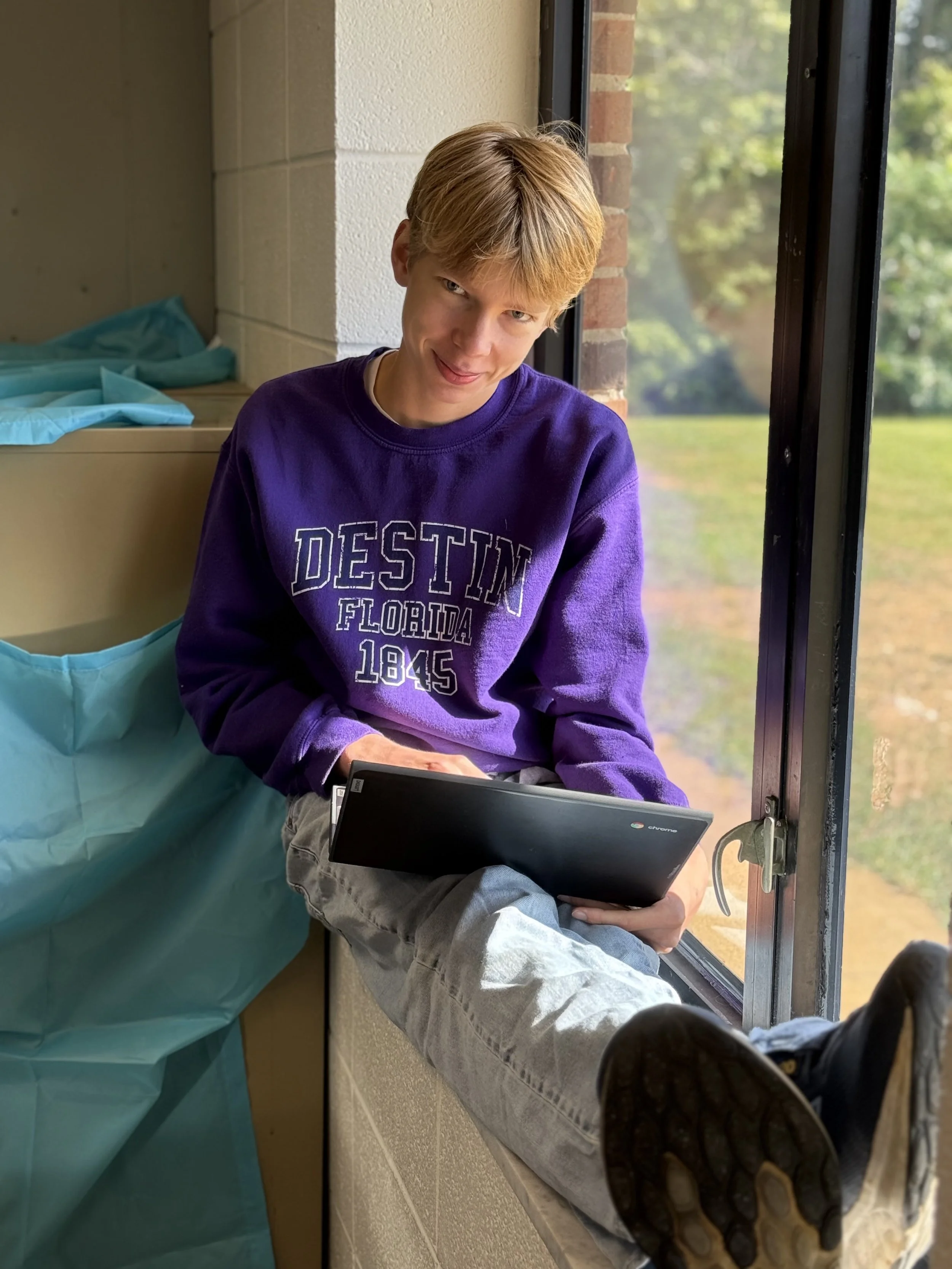 A teenage boy with blond hair sitting on a windowsill, holding a Chromebook in his lap, wearing a purple sweatshirt that says 'DESTIN FLORIDA 1845,' looking at the camera with a slight smile.