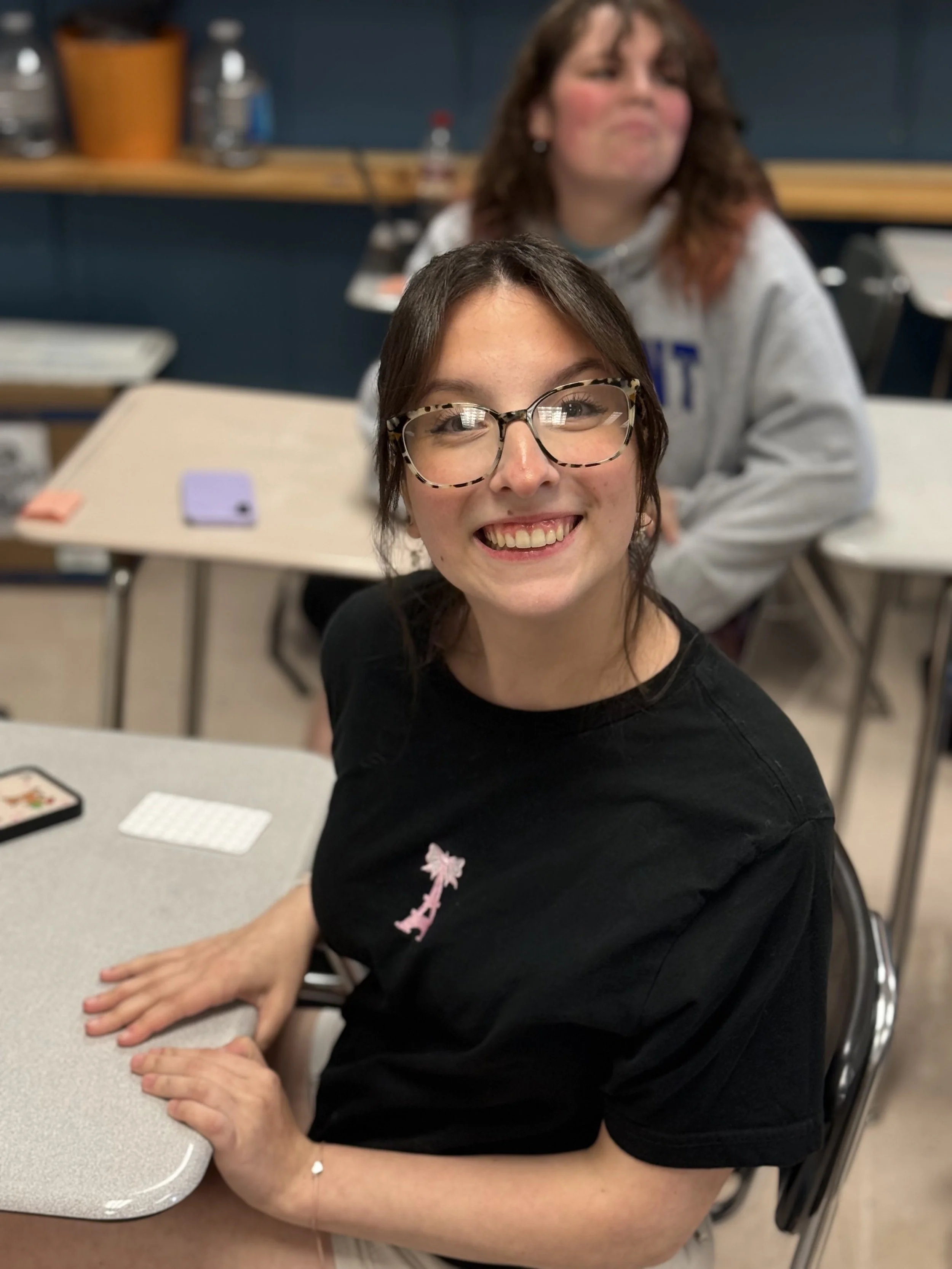 Smiling young woman with glasses and dark hair, sitting at a desk in a classroom, with another person blurred in the background.
