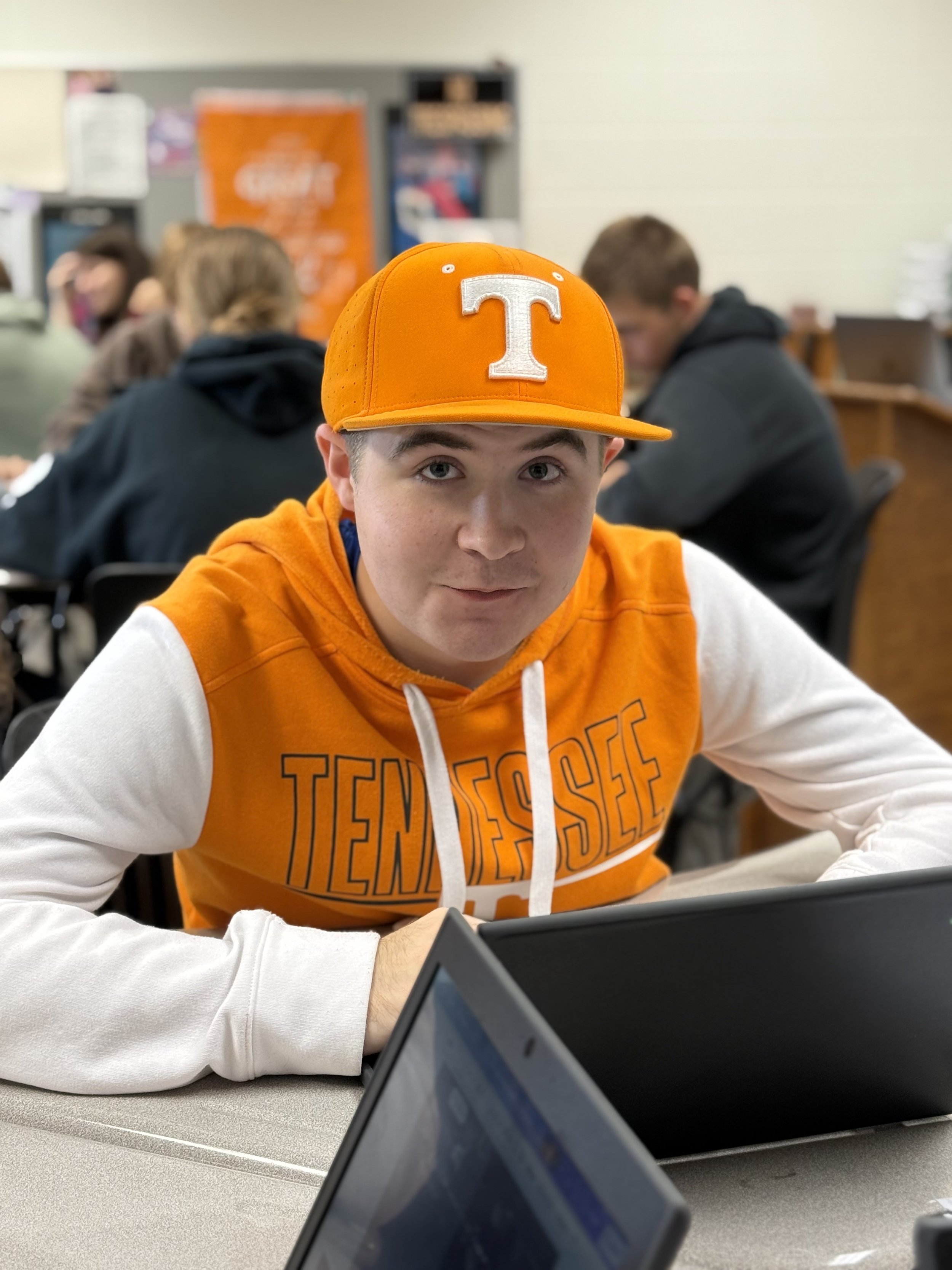 A young man wearing an orange Tennessee Volunteers hoodie and cap, sitting at a desk in a classroom or café, with several people in the background.