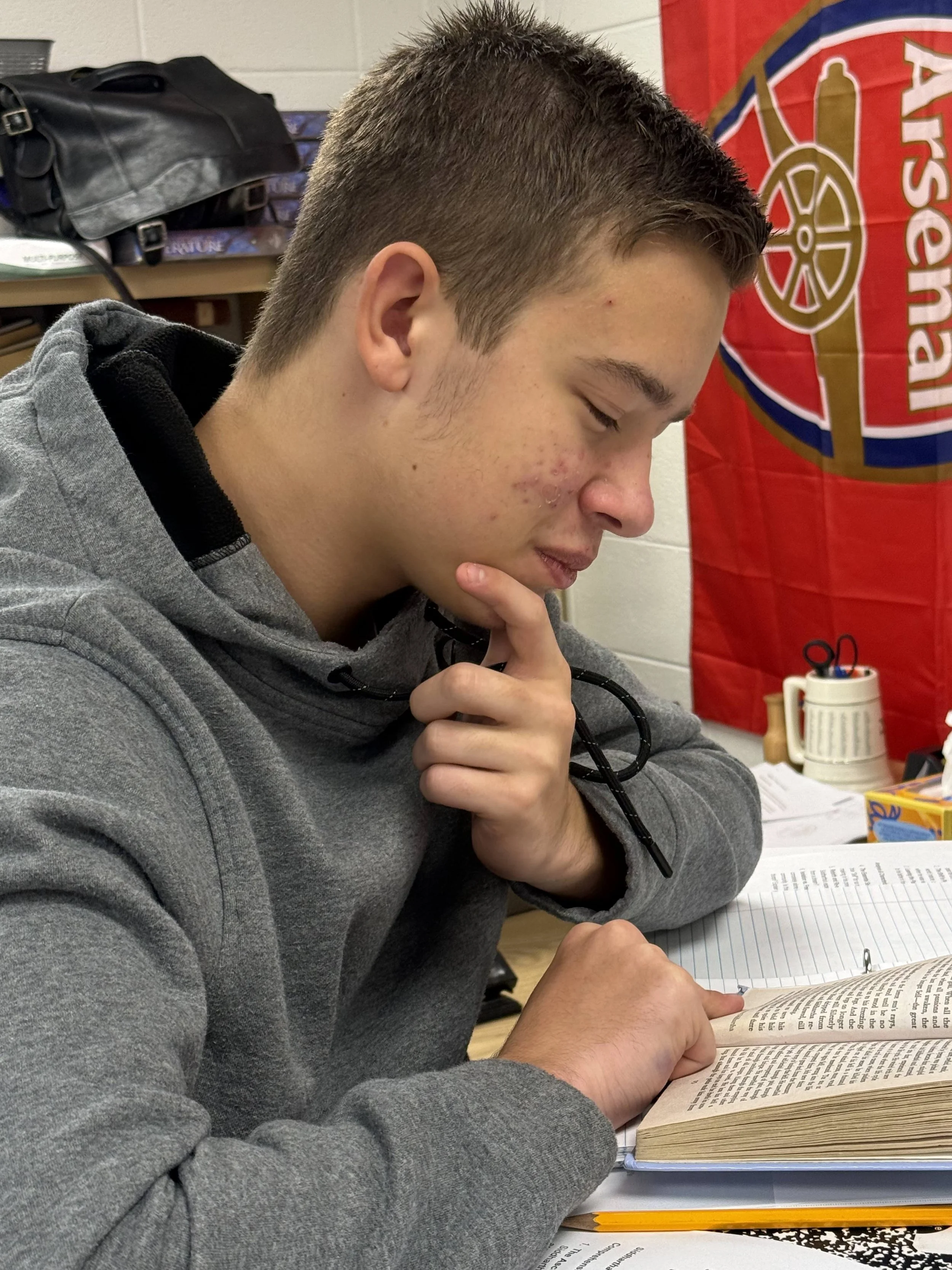 A young boy sitting at a table giving a thumbs-up, with a classroom bulletin board behind him. The bulletin board has various colorful posters and papers, including one with a rainbow and the words "Be kind" and others with text about different topics. The table has a green notebook, a laptop, and a bottle of water.