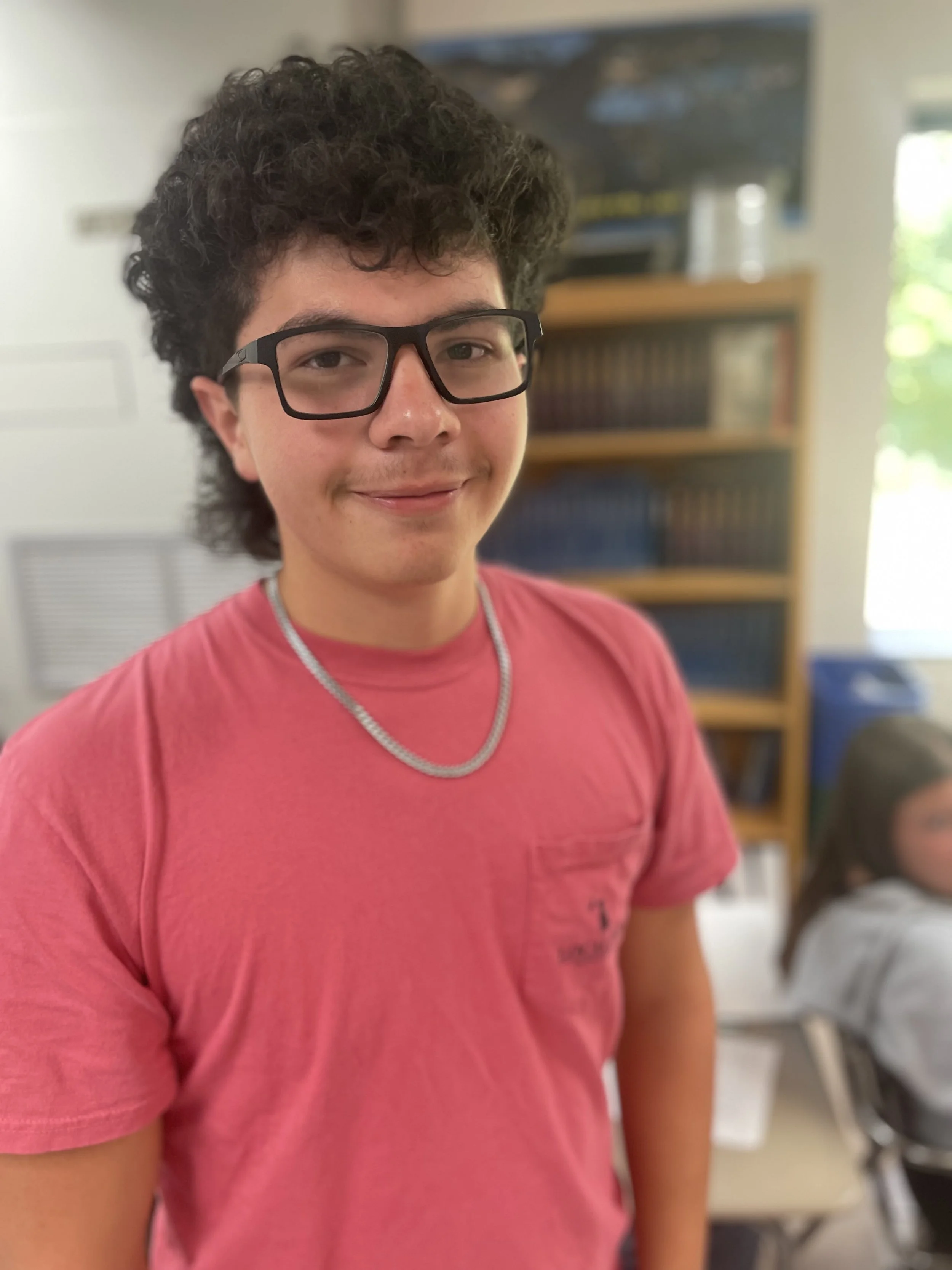 A young man with curly dark hair, wearing glasses, a pink T-shirt, and a silver chain necklace, smiling in a classroom.