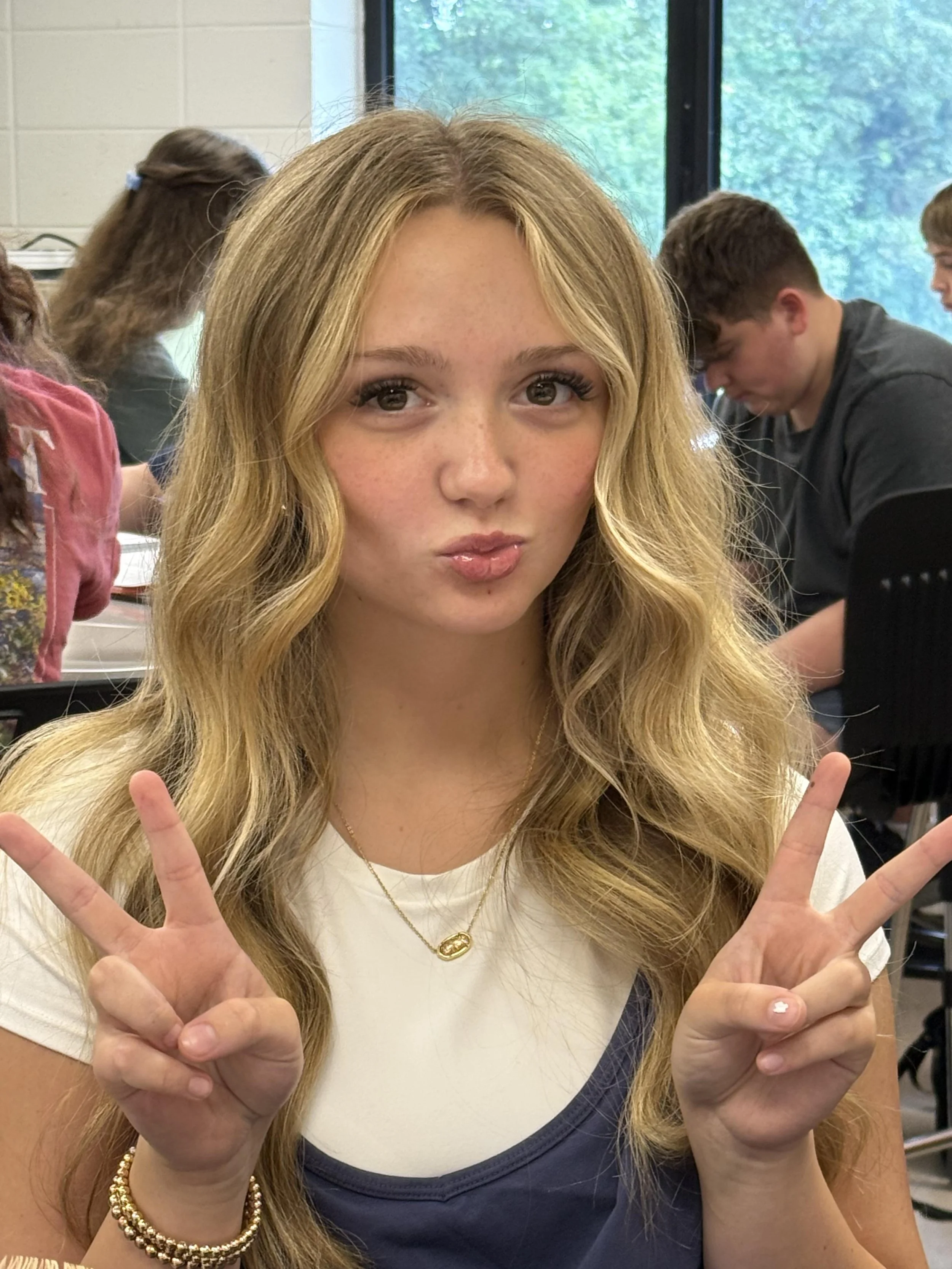 A young woman with long wavy hair and freckles, smiling at the camera, sitting in a classroom with a whiteboard and shelves of binders behind her.