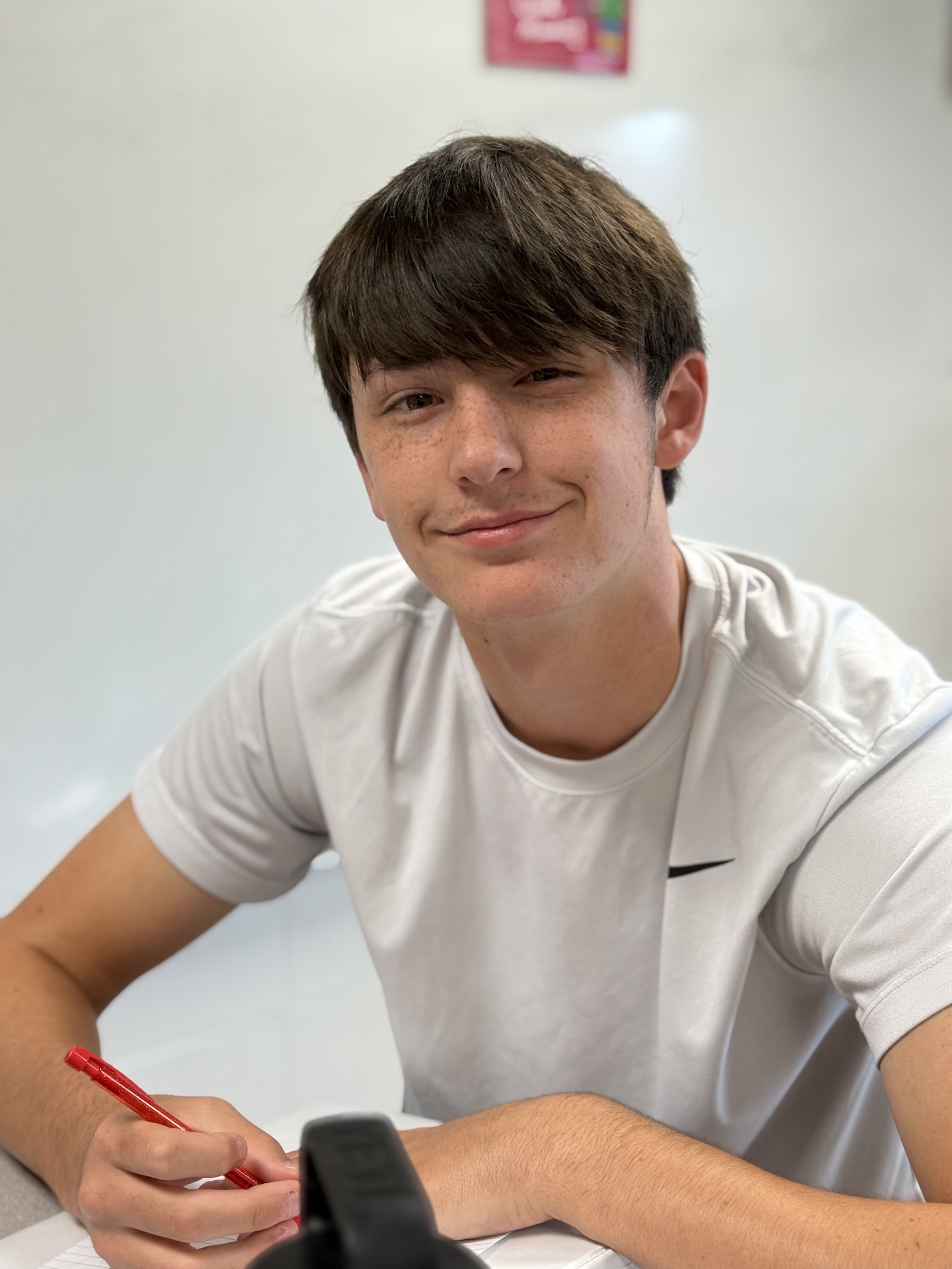 Young man with brown hair and freckles smiling at the camera, wearing a white t-shirt, sitting at a table with a red pen and a notepad.