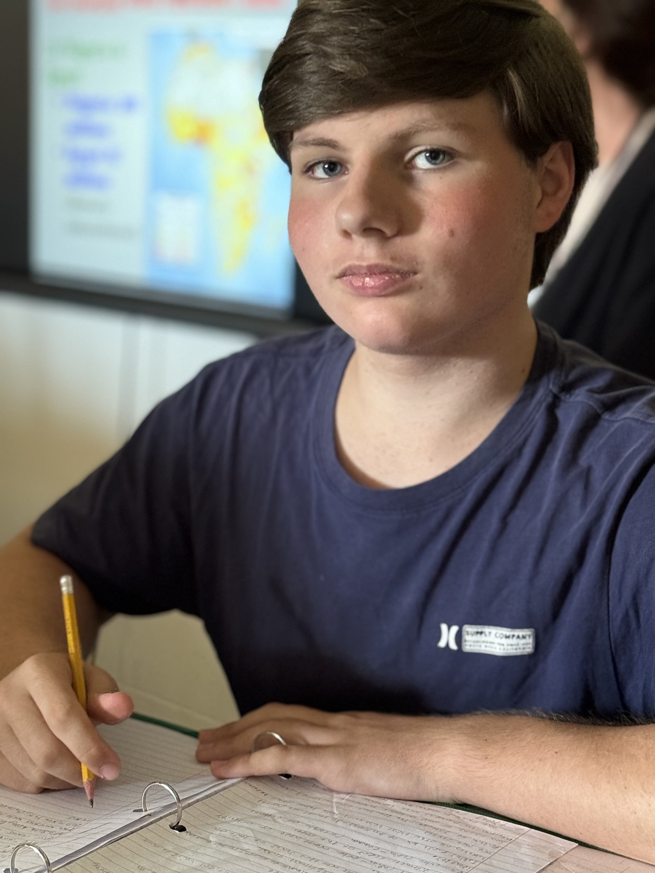 A teenage boy with brown hair and blue eyes is sitting at a desk in a classroom, holding a yellow pencil, with a notebook open in front of him. There is a blurred classroom presentation in the background.
