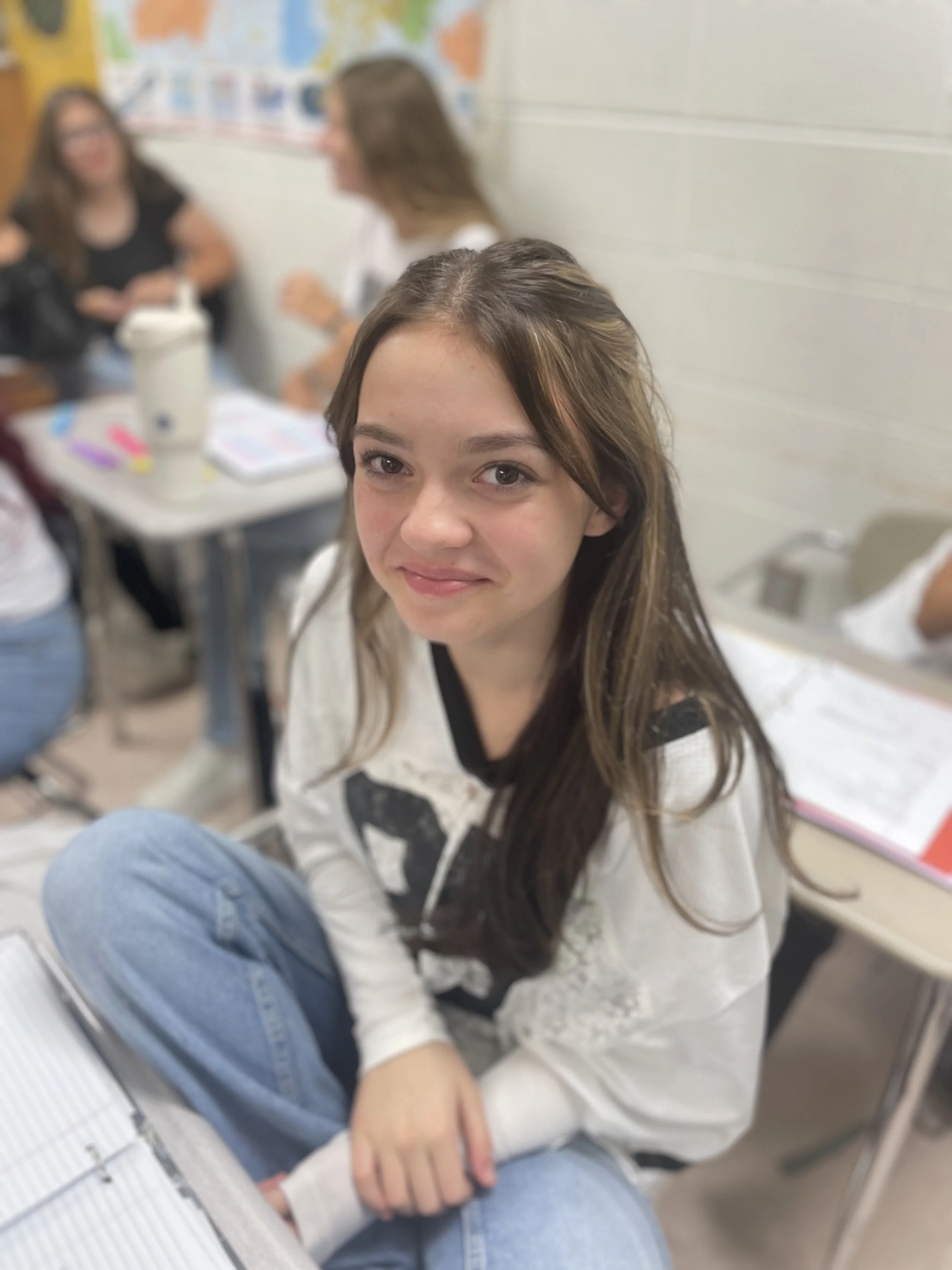 A young girl with long brown hair and a white shirt, sitting cross-legged at a desk in a classroom, smiling at the camera.