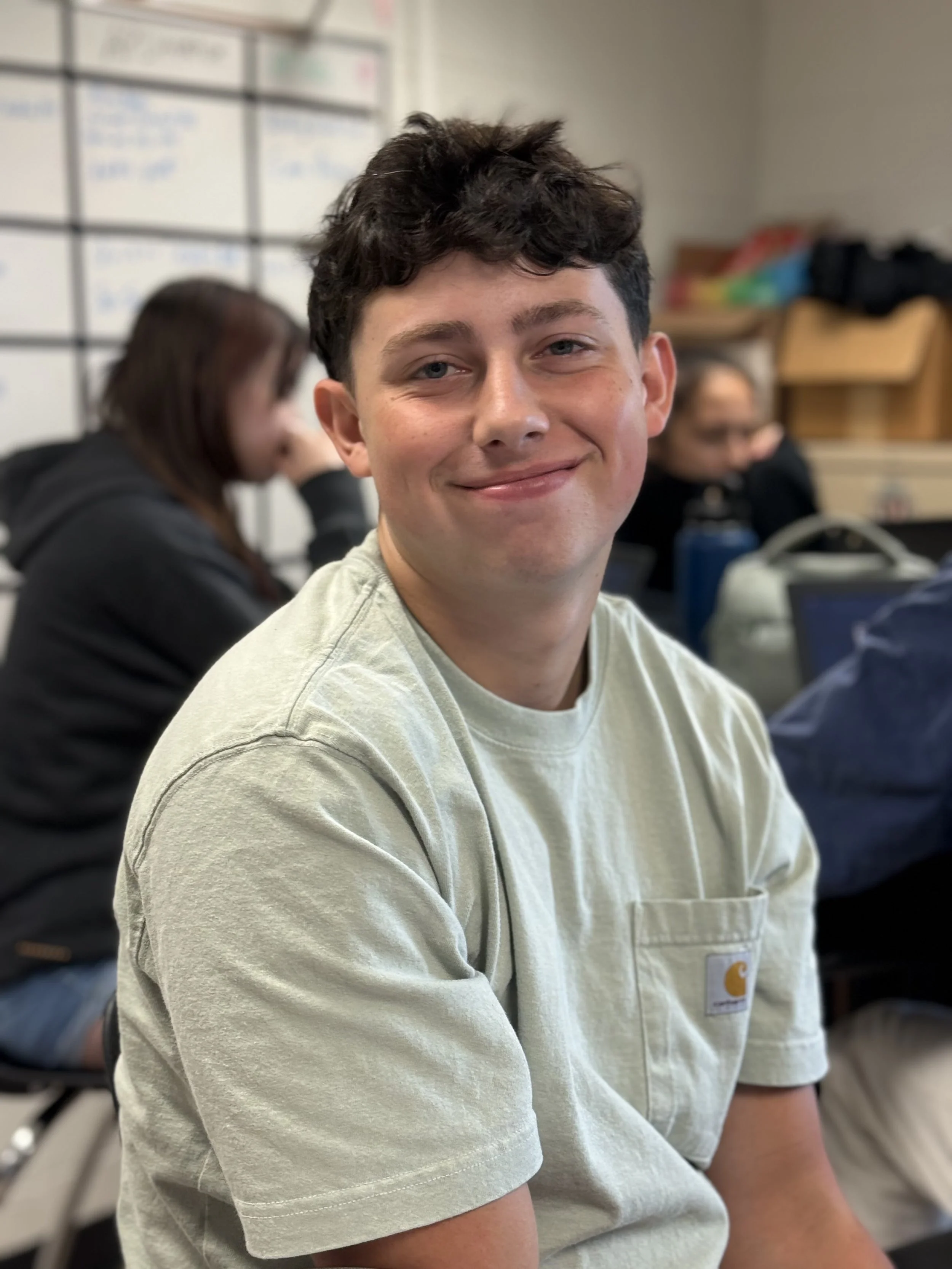 A young man smiling in a classroom with other students blurred in the background.