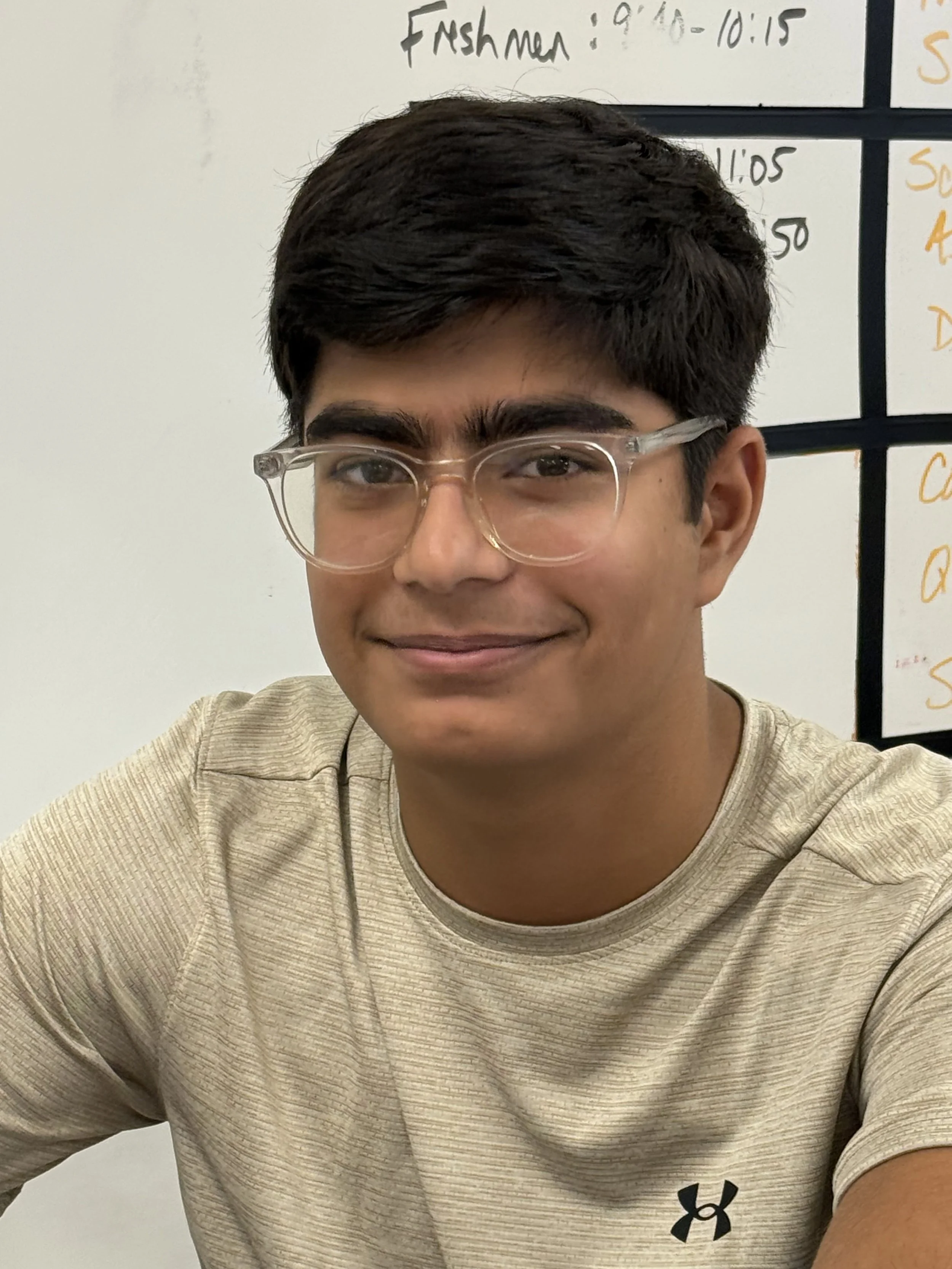 A young man with glasses smiling, sitting at a desk in a classroom with other students in the background.